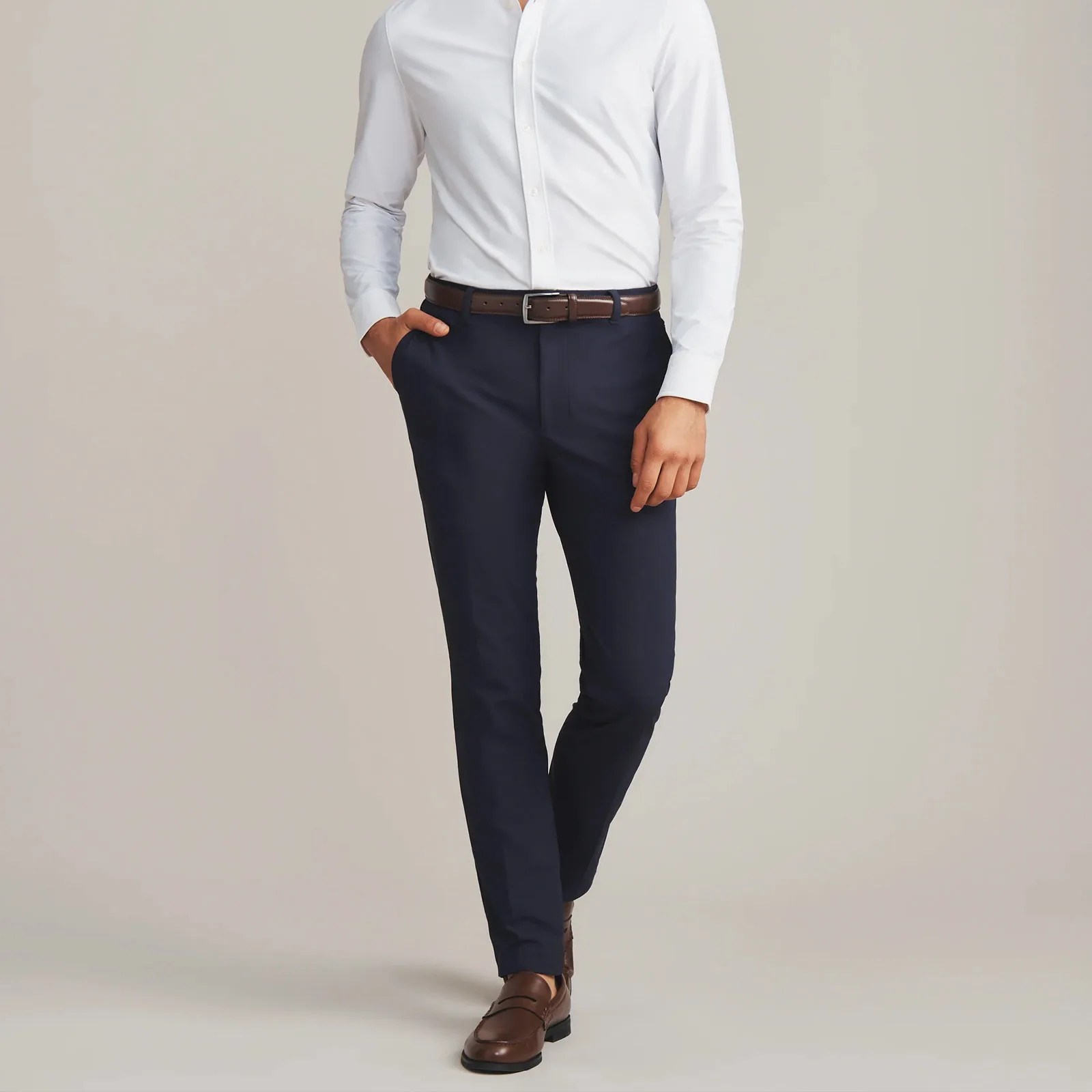A male model wearing a navy pair of Rhone Commuter Pro Pants shown from the shoulders down facing forward while taking a step. The model's hand is sitting in one pant pocket. The model is also wearing a white button dress shirt, brown belt, and brown shoes. 