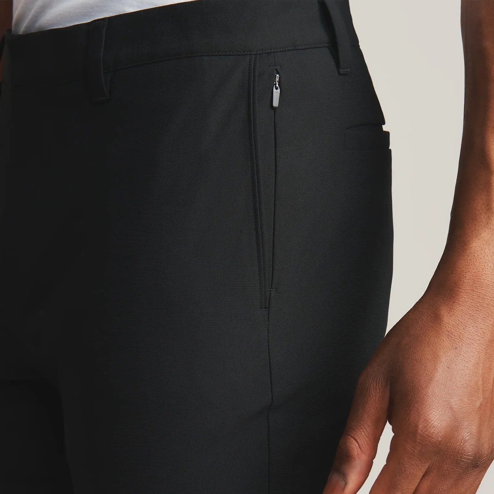 A close-up view of the zipper pocket of the Rhone Commuter Pant Pro in Black is worn by a dark-skinned model. Only the left hip and a silver of the model's forearm and hand are visible and in frame. 
