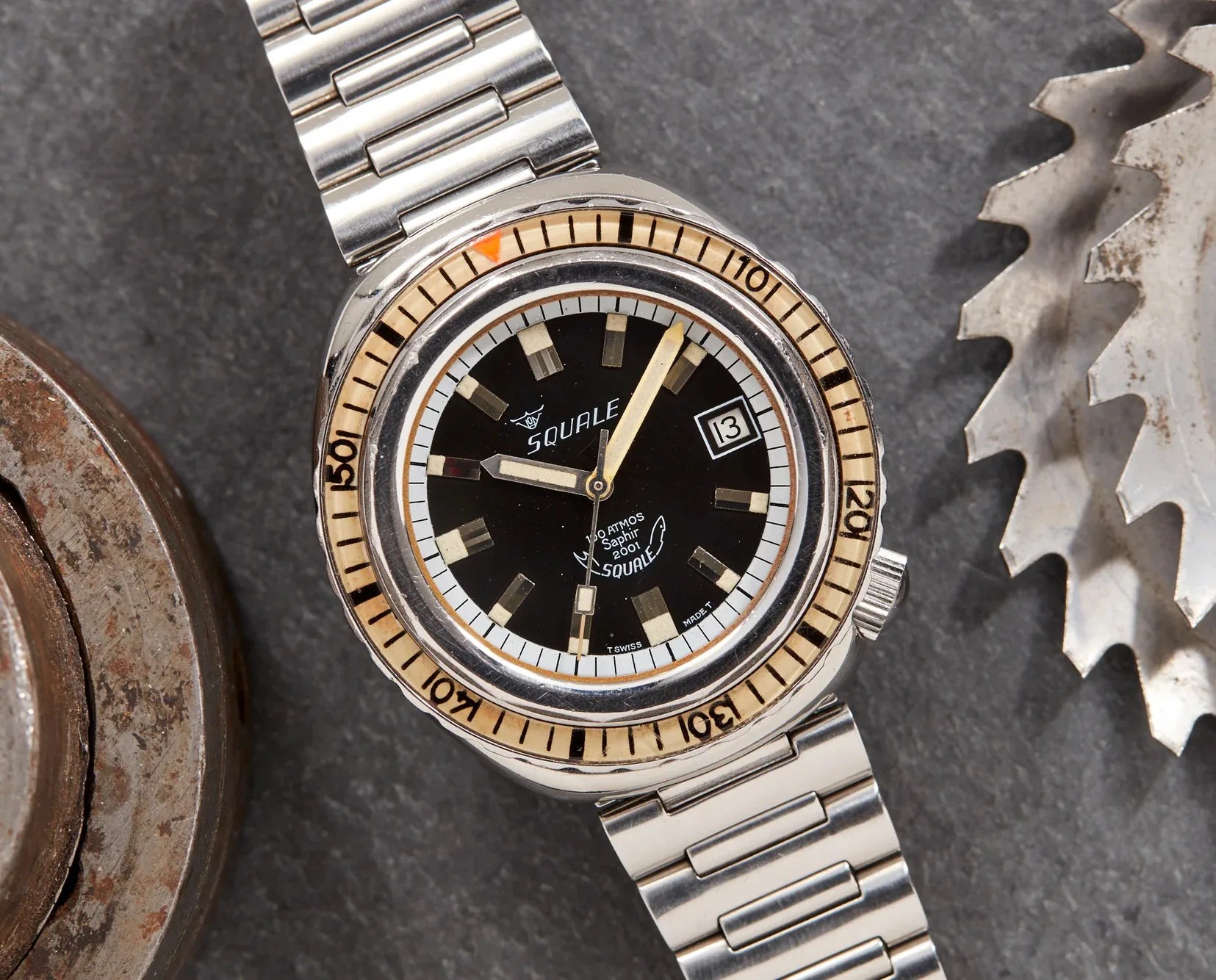 A Squale Saphire 2001 vintage dive watch photographed from above positioned between what appears to be a a circular metallic saw black and metallic grinder.