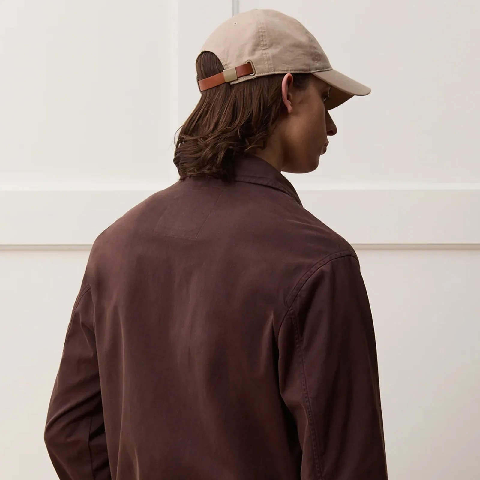 A male model with shoulder length brown hair wearing a light tan dad cap made by Todd Snyder shown from behind. The leather strap of the hat is visible. the model is also wearing a chocolate brown jacket. 