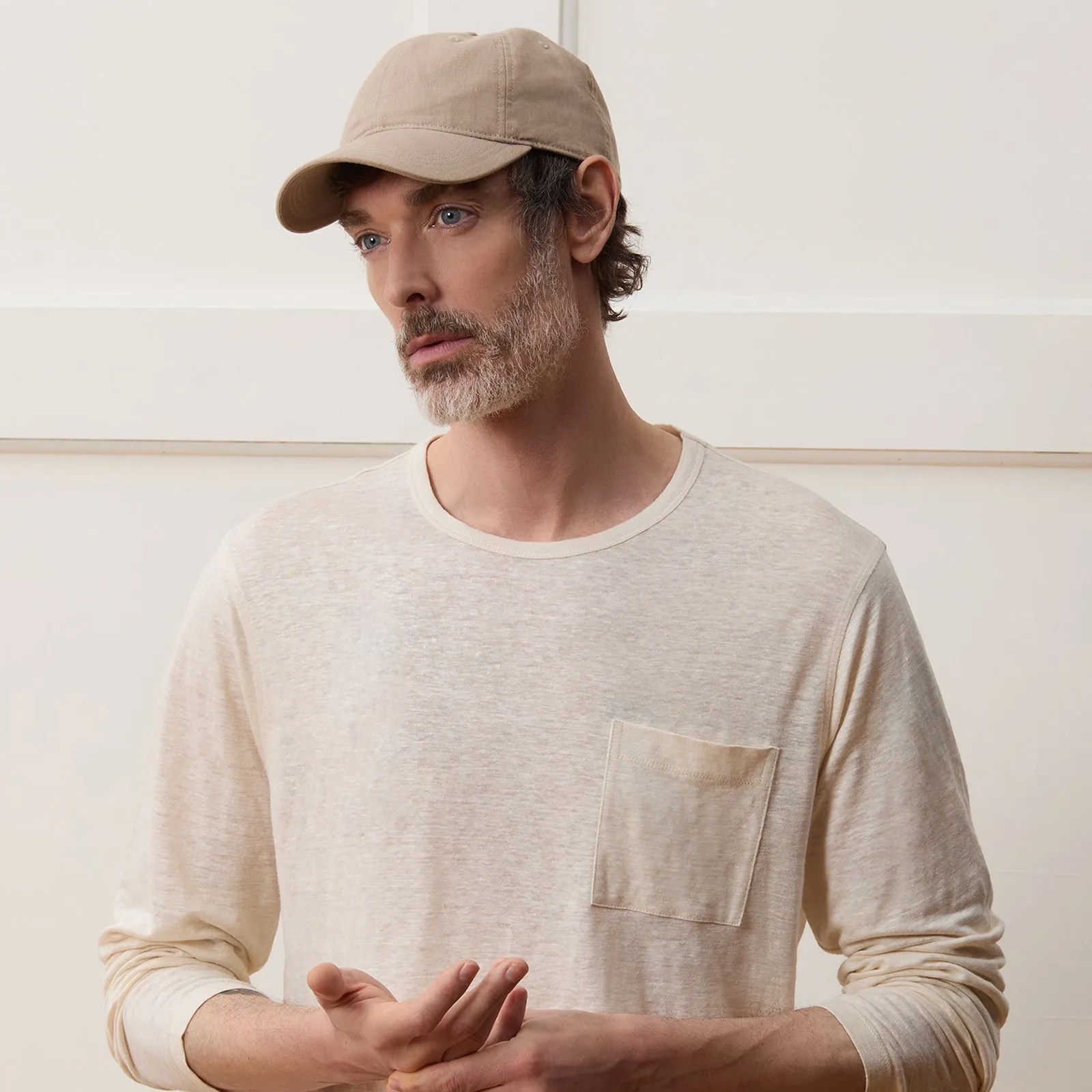A man wearing a Todd Snyder Linen Dad Cap in light tan. The man is shown at an angle looking to the left of the image frame. He is wearing a creamed color long sleeve t-shirt with a pocket and standing in front of a cream-colored wood paneled wall. 