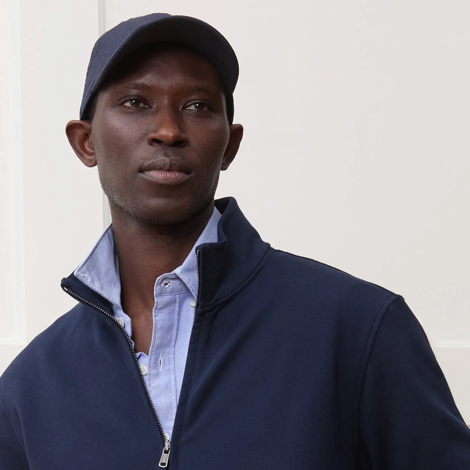 A black male model wearing a navy quarter zip jacket, blue button up shirt and navy dad cap all made by Todd Snyder. The model is facing forward with his eyes looking out and to the right of the image frame. The jacket is partially unzipped to reveal the light blue shirt underneath. The model is shown from the mid waist up. 