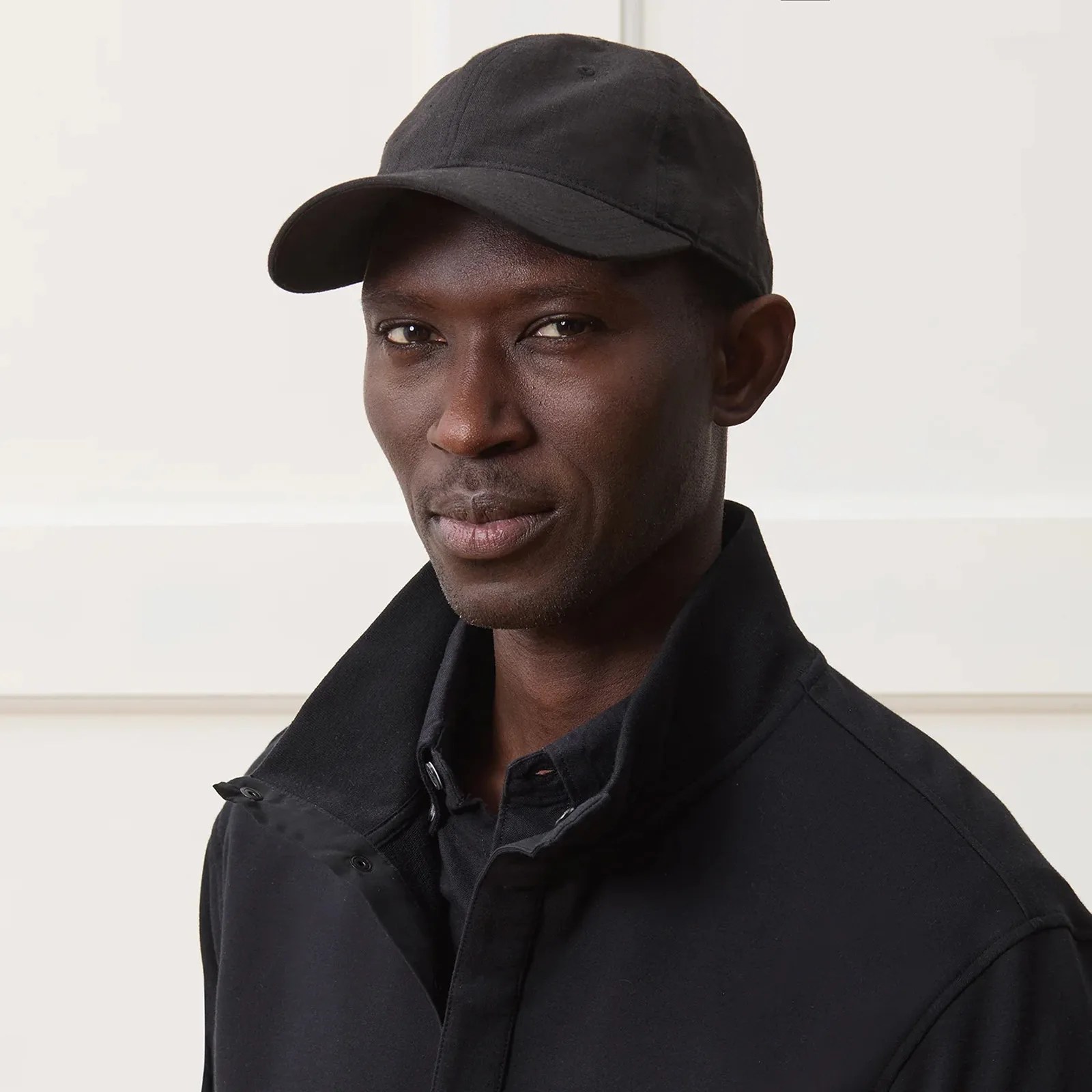 A black male model shown from the chest up facing forward wearing a black Todd Snyder designed dad hatt and black jacket. Behind him is a cream white paneled wood wall 