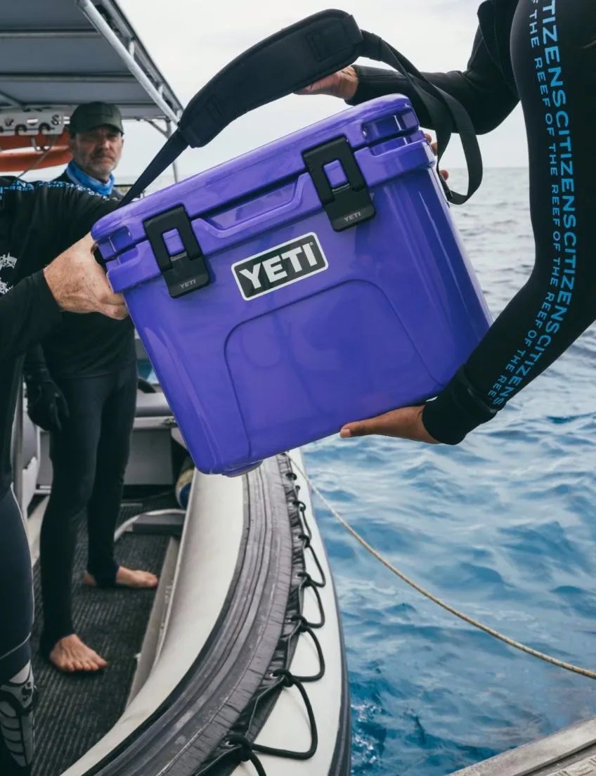 A small Yeti hard cooler in Ultramarine purple being handed to a person standing on a dive boat wearing a wetsuit. The person handing the cooler is also wearing a wetsuit and standing on another boat.
