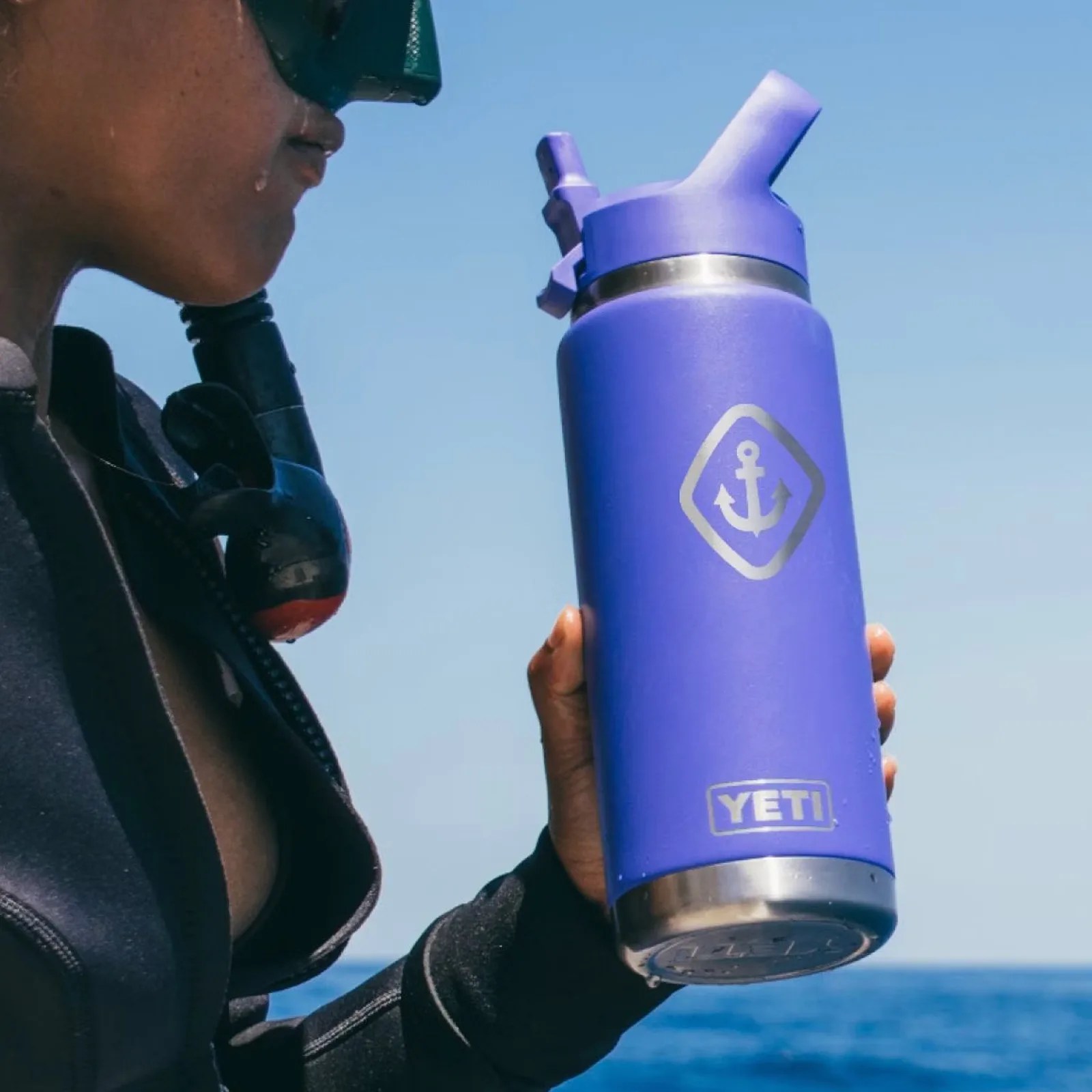 A woman in snorkling gear who appears to have just gotten out of the water shown profile holding up a Yeti Rambler water bottle in Ultramarine which is a purplish blue hue. The ocean can be seen int he background. 