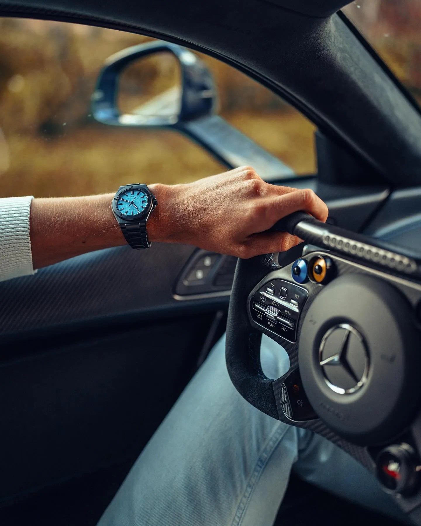 F1 Driver George Russell's wrist is shown holding a Mercedes supercar steering wheel, showing his custom Ingenieur Automatic 42 in black ceramic with a custom blue face. 