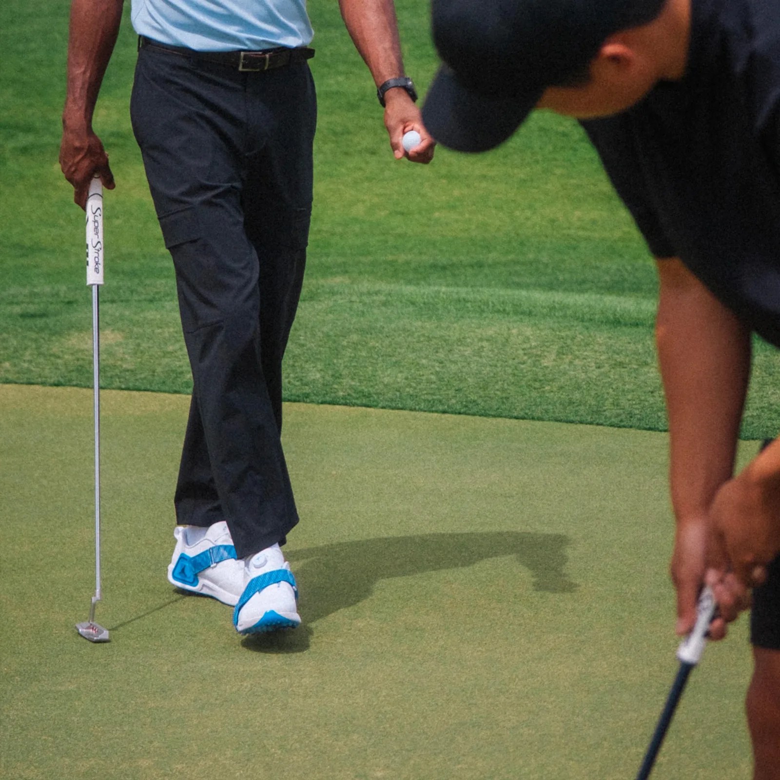 A golfer on a green is walking towards another golfer, putting shown from the waist down. The golfer is wearing a pair of Jordan Brand Air Rev golf shoes in white with bright blue accent straps.