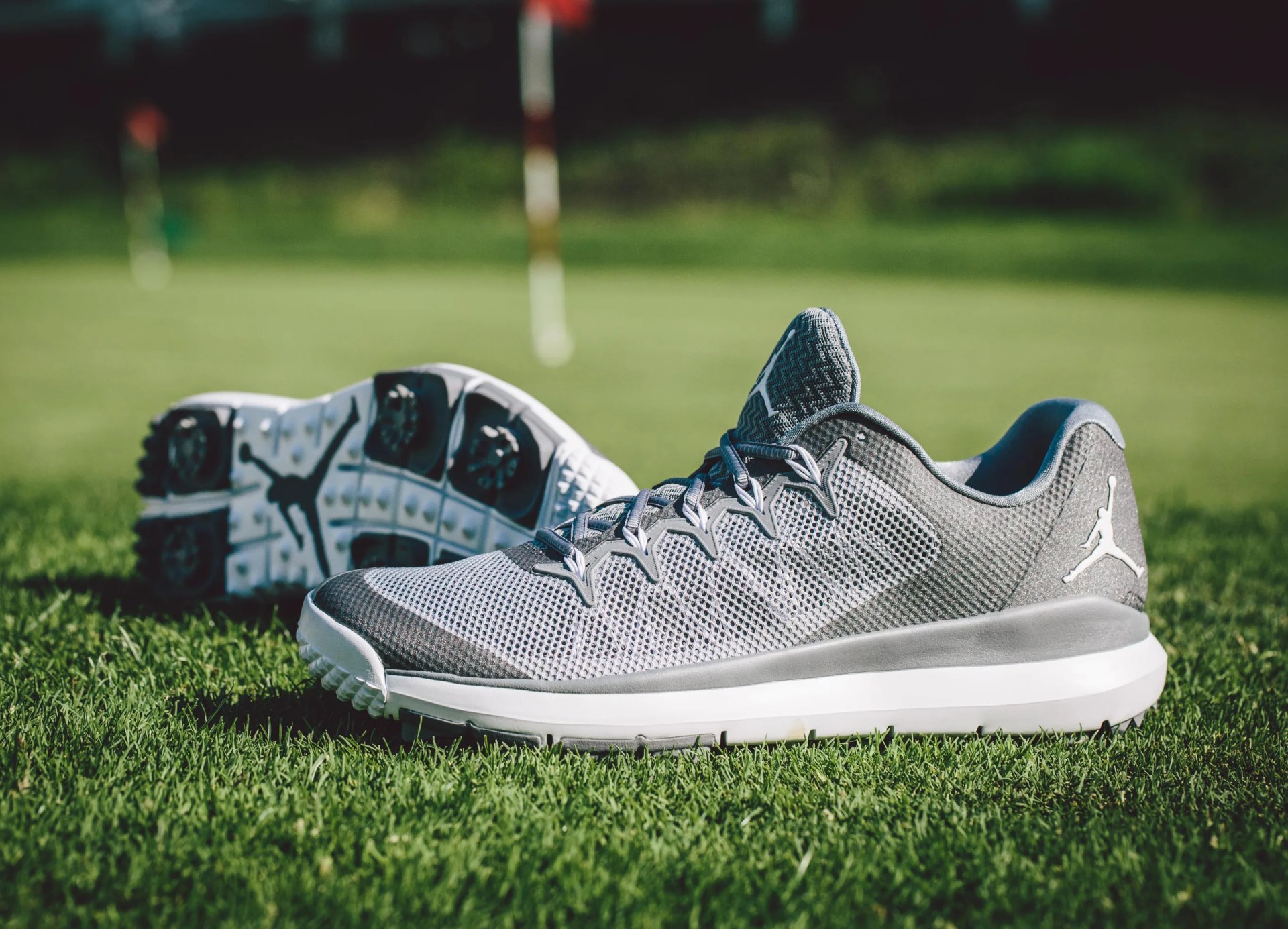 A pair of grey and white Jordan Flight Runner 2 golf shoes is shown sitting on a golf course green with a golf flag blurred in the background. One shoe is shown sitting upright in profile with the toe facing to the left. Another is shown in the background lying diagonally on its side to reveal the spiked outsole. 
