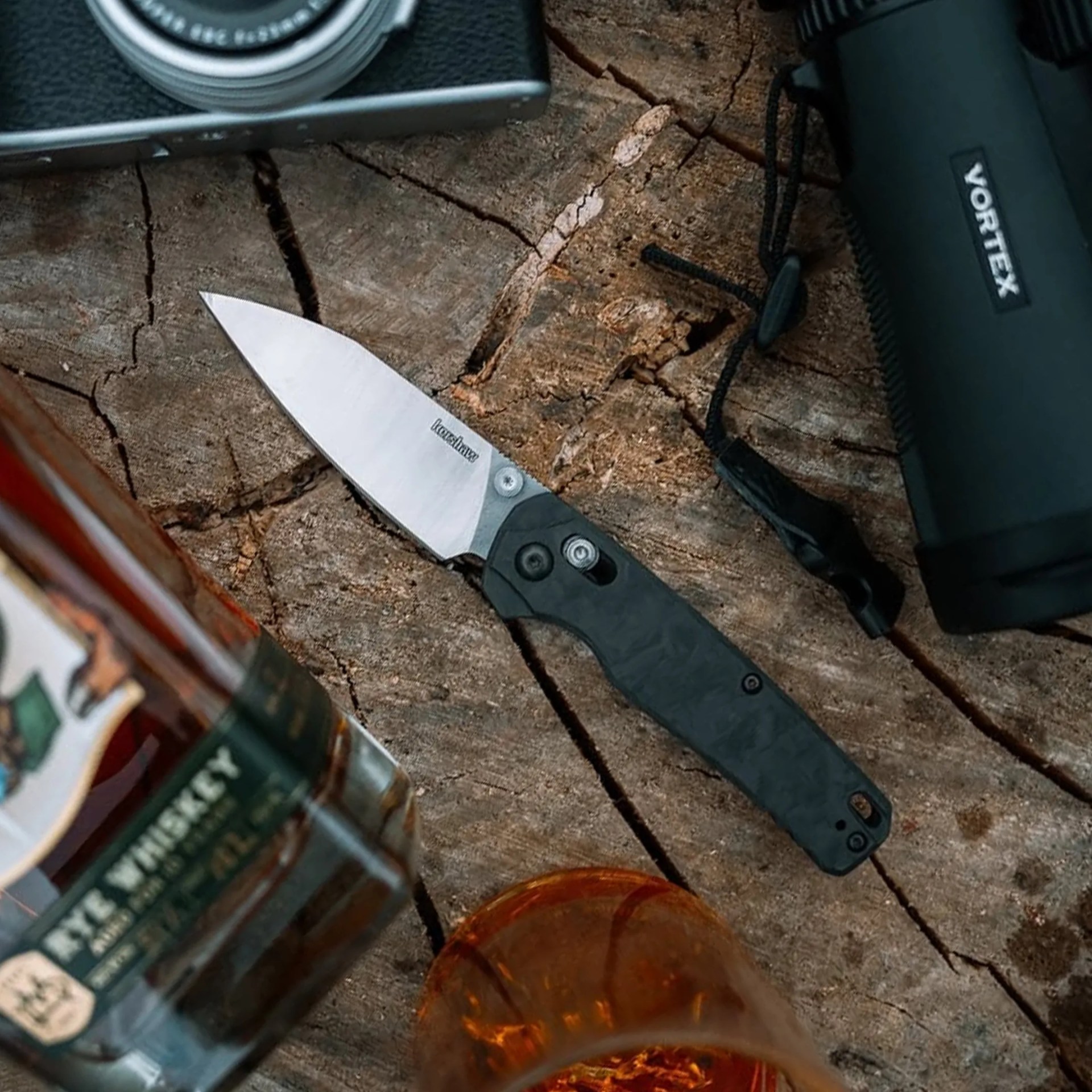 Kershaw Bel Air Carbon Fiber EDC Knife on stump next to camera, binoculars, whiskey