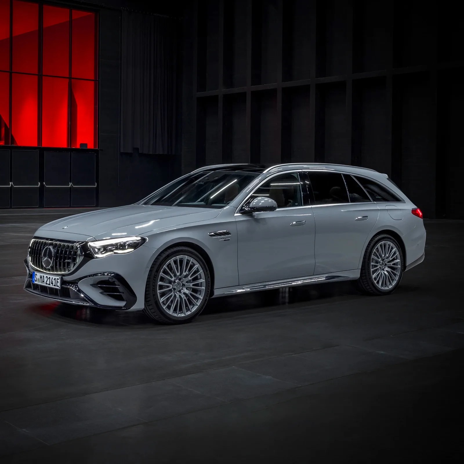 The 2026 Mercedes-AMG E53 Hybrid Wagon is shown from the side in grey in a rendered black warehouse. 
