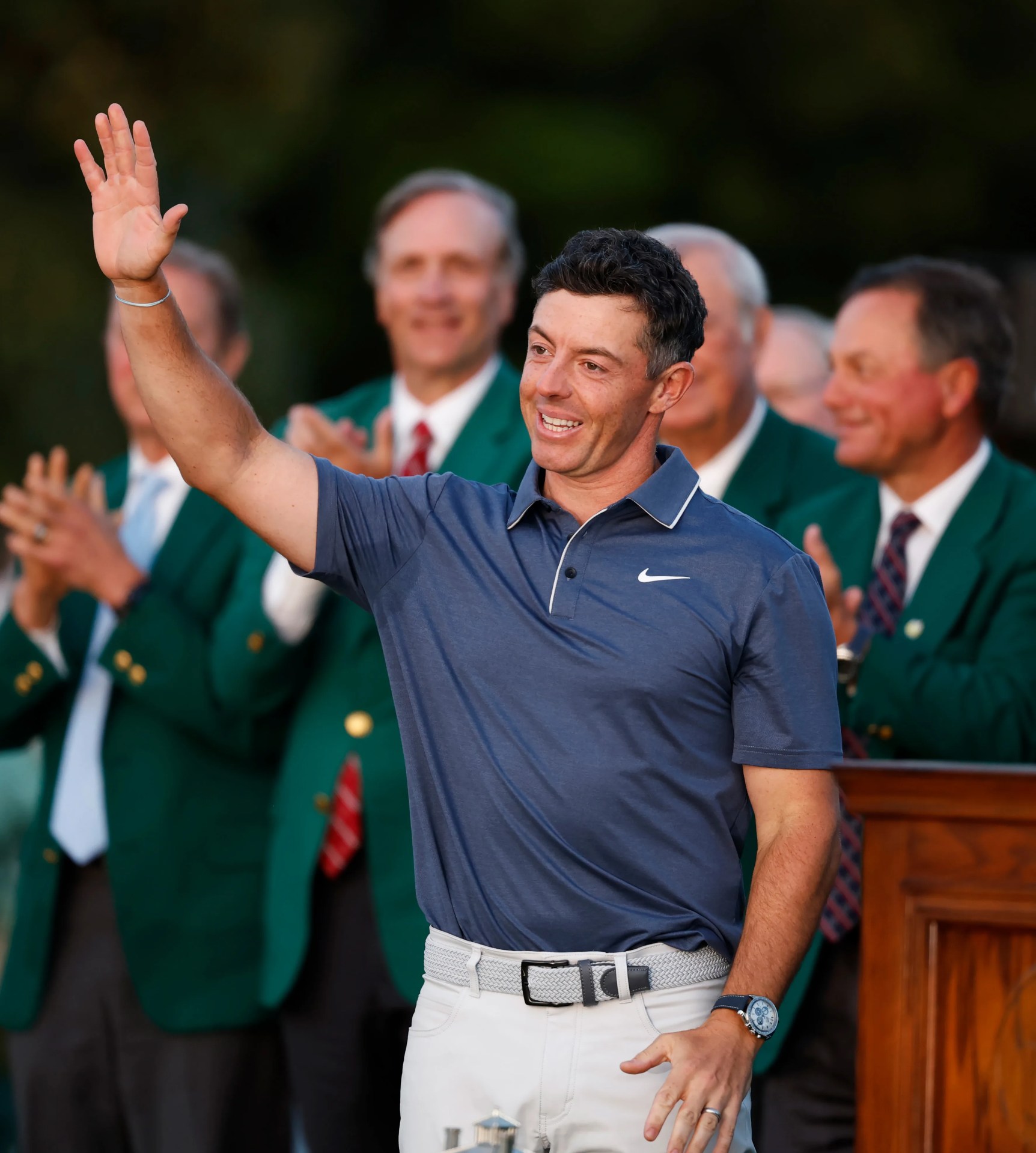 Rory McIlroy is shown with his hand in the air waving at the 2025 Masters Green Jacket Ceremony. Rory is wearing a navy nike golf shirt, white pants an Omega Speedmaster "Silver Snoopy Award" 50th Anniversary Series watch on his wrist. Behind him are men blurred in the background wearing green jackets.