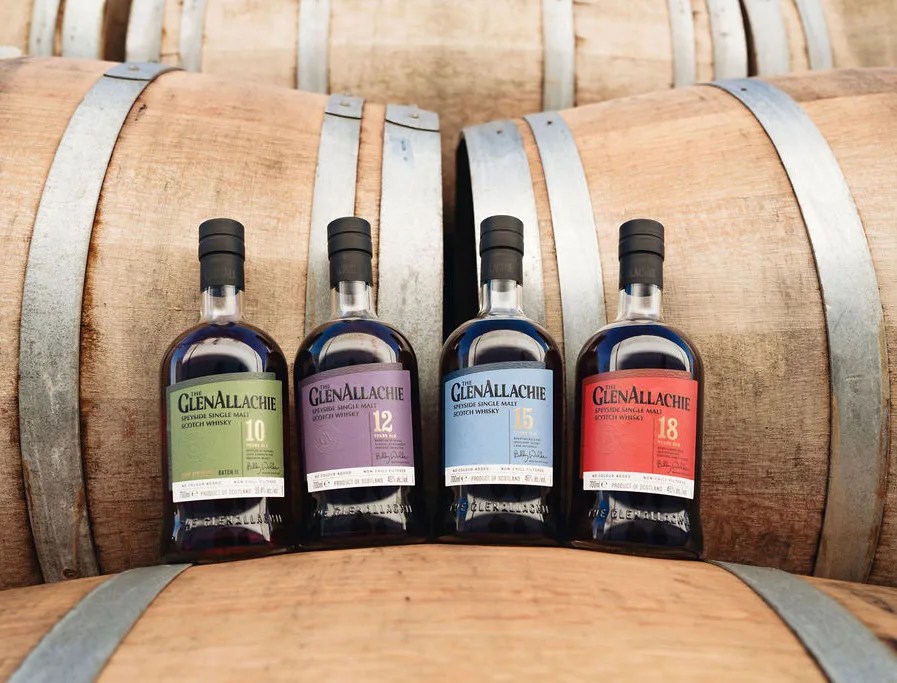 Four bottles of The GlenAllachie whisky core collection shown in a row sitting on whisky barrels. 