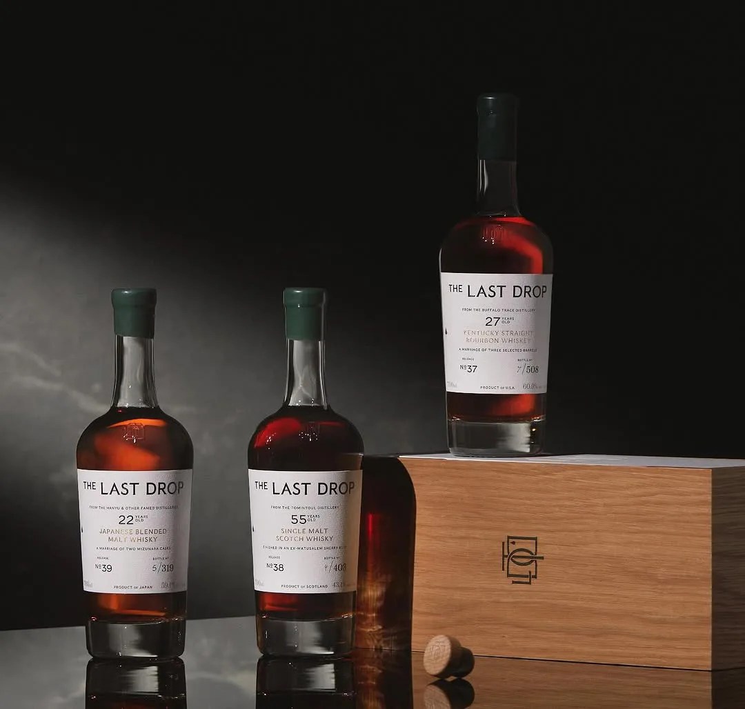 A collection of three bottles of The Last Drop whiskies. Two are sitting on a shiny table, and one is sitting on top of a wooden whiskey box with the Last Drop emblem. 
