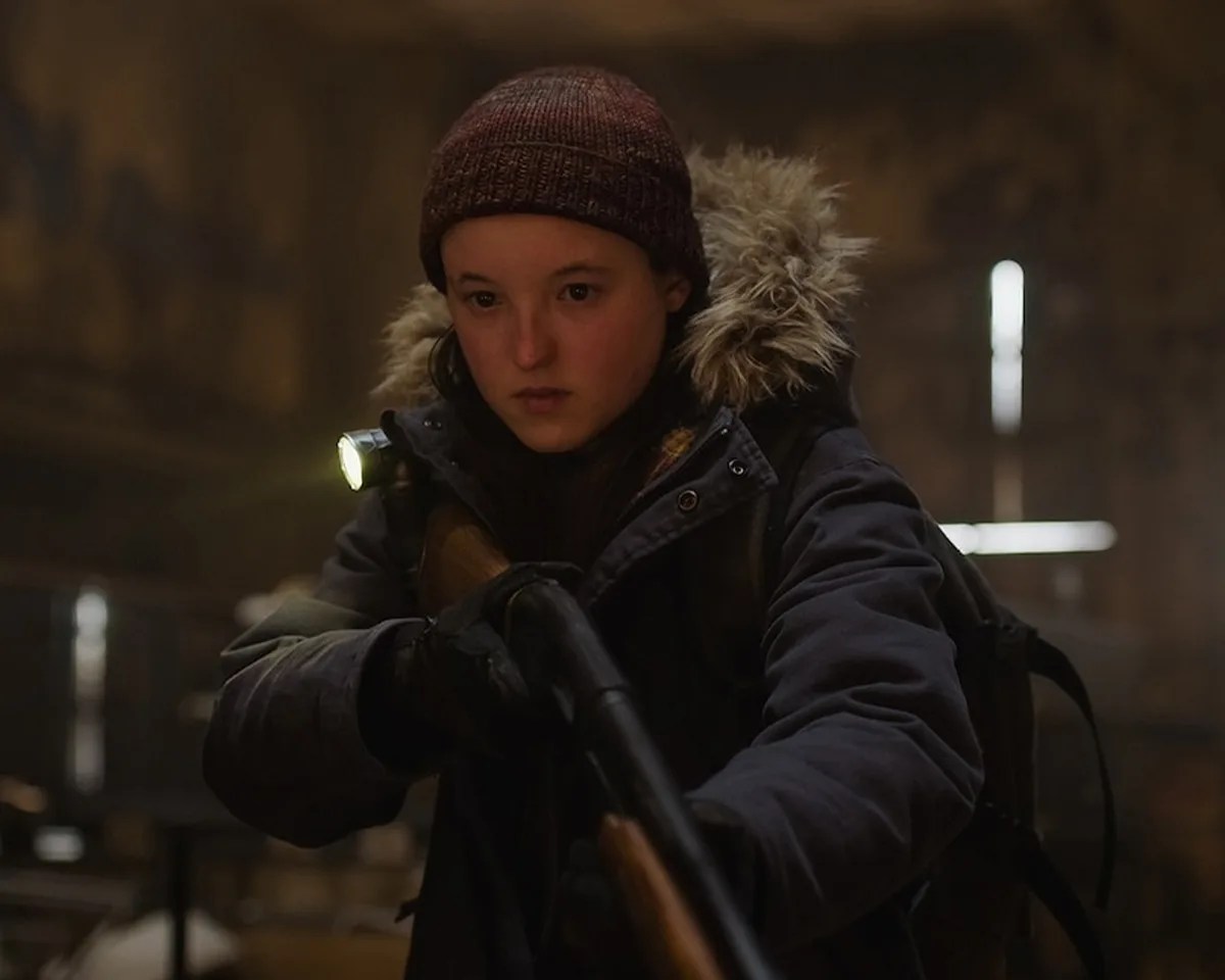 Bella Ramsay as Ellie in beanie and fur-lined jacket holding shotgun
