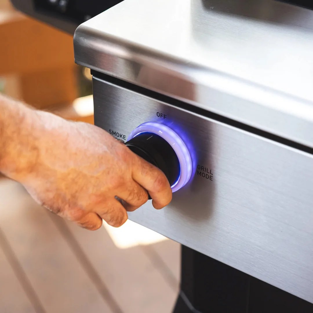 A hand shown adjusting the cooking control knob that switches between smoke and grill mode on the Recteq X-Fire Pro 825 pellet grill. The knob is shown illuminated with a purple ring. 