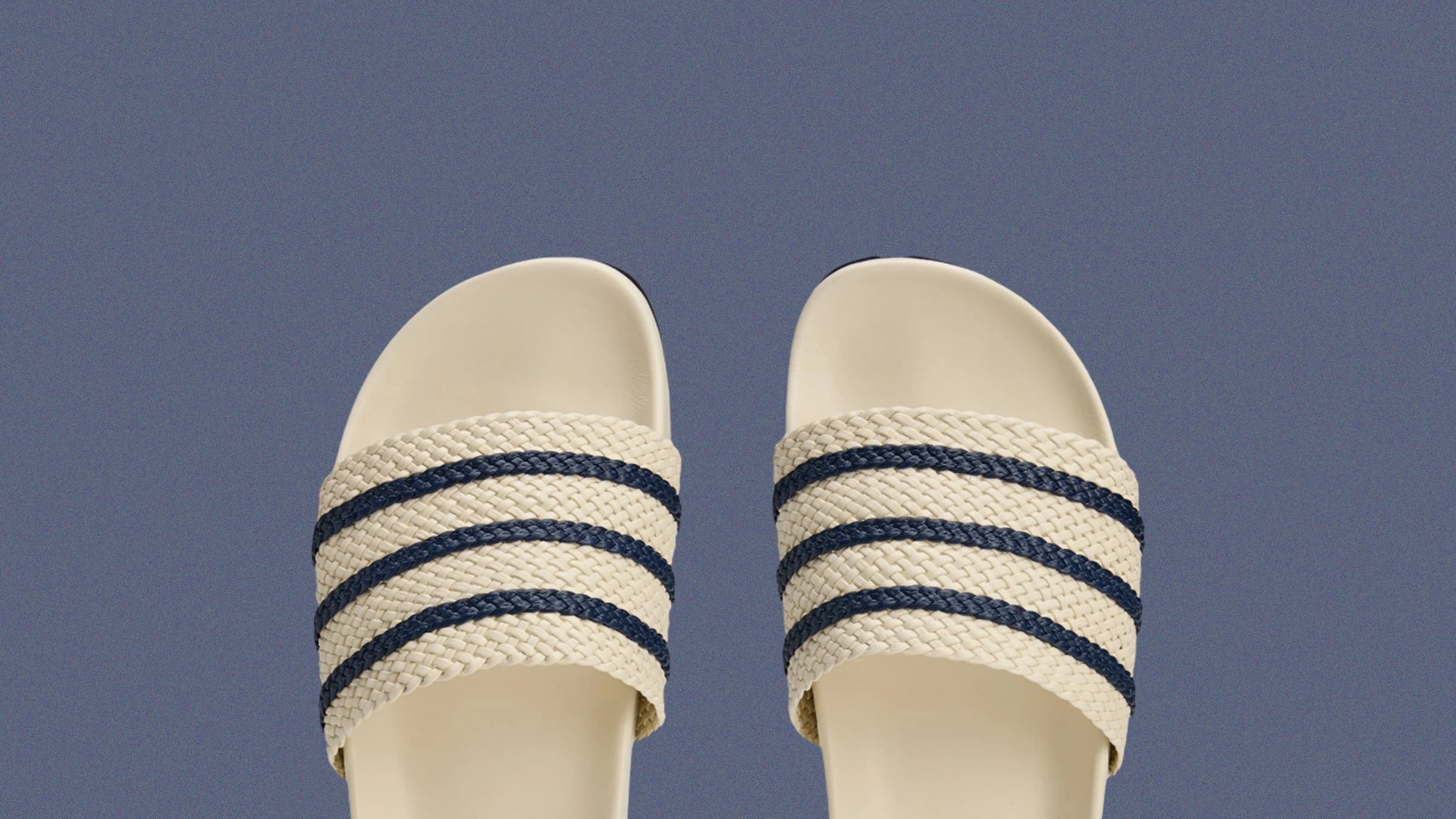 A pair of Wale Bonner Adidas Adilette recovery sandals in cream white with blue strips shown peeking out from the bottom edge of an image with a speckled blue background