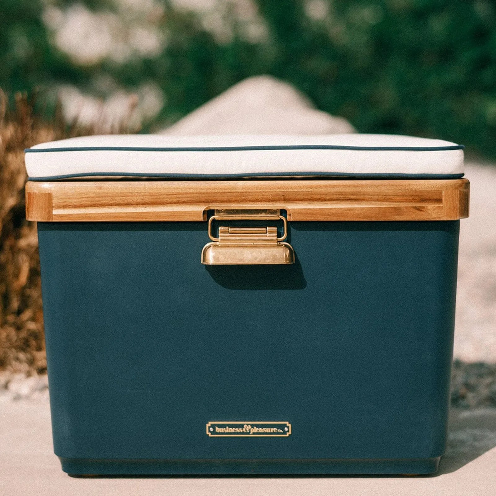 A navy blue colored Business&Pleasure Co. The Hemingway Cooler sitting on a beach with a blurred background.
