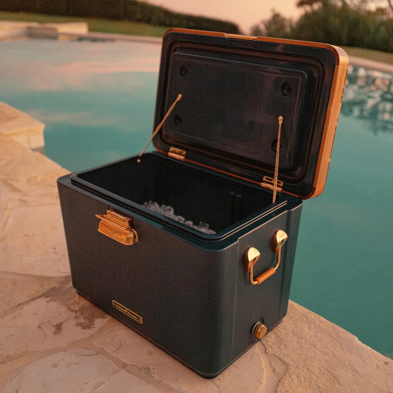 The Business & Pleasure Co.The Hemingway Cooler in Navh is shown with the lid open to reveal ice on the inside. The cooler is sitting on the edge of a swimming pool. 