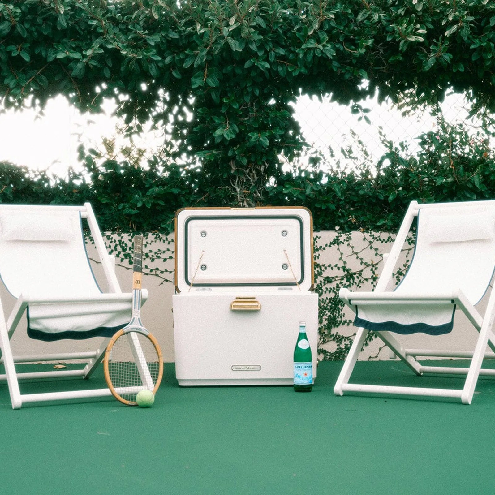 The Hemingway Cooler by Business & Pleausre Co. in white with the lid open positioned between two white outdoor reclining chairs on a tennis court surface