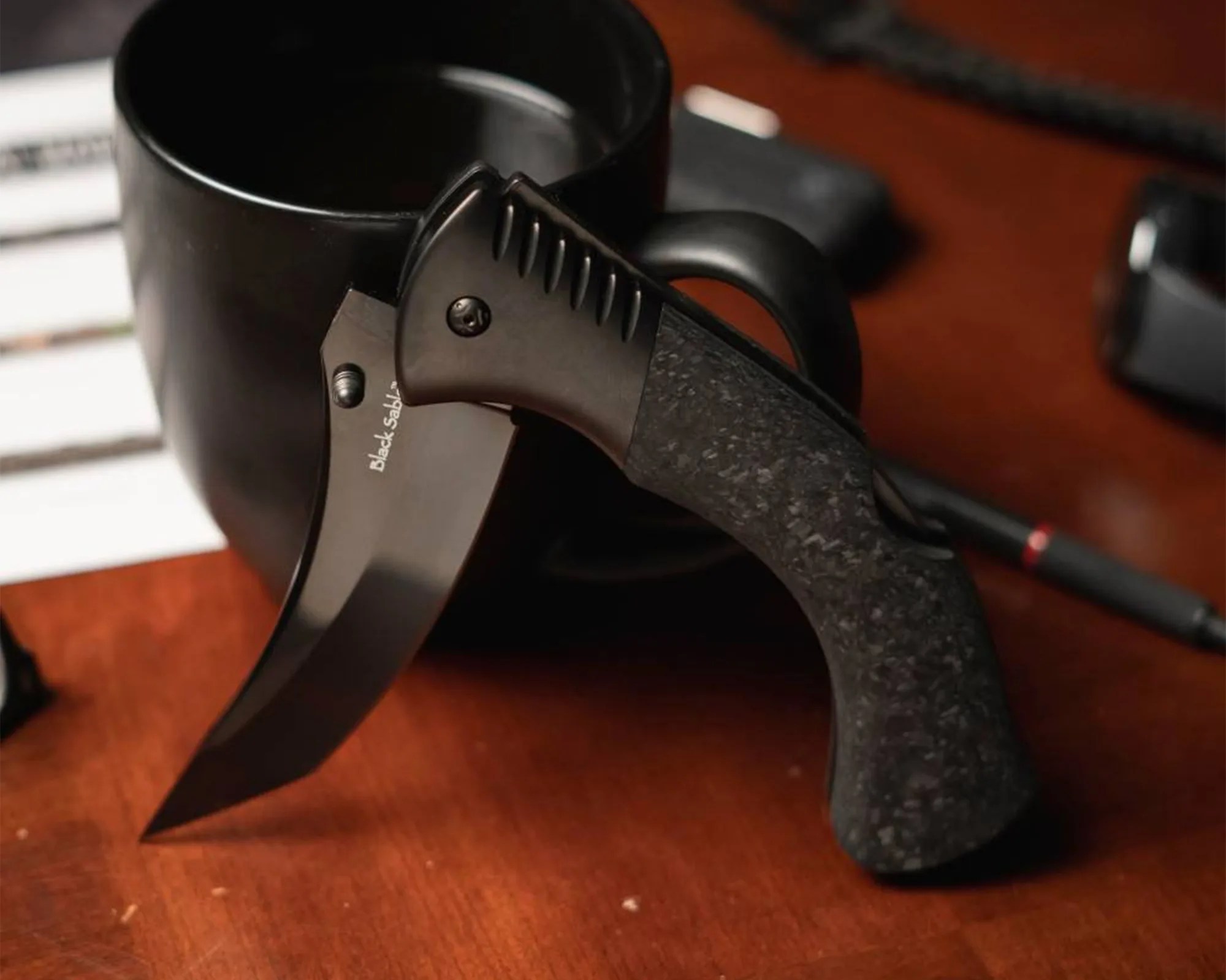 Cold Steel Black Sable EDC Knife on coffee mug on wood desk with watch, glasses, pen, etc.