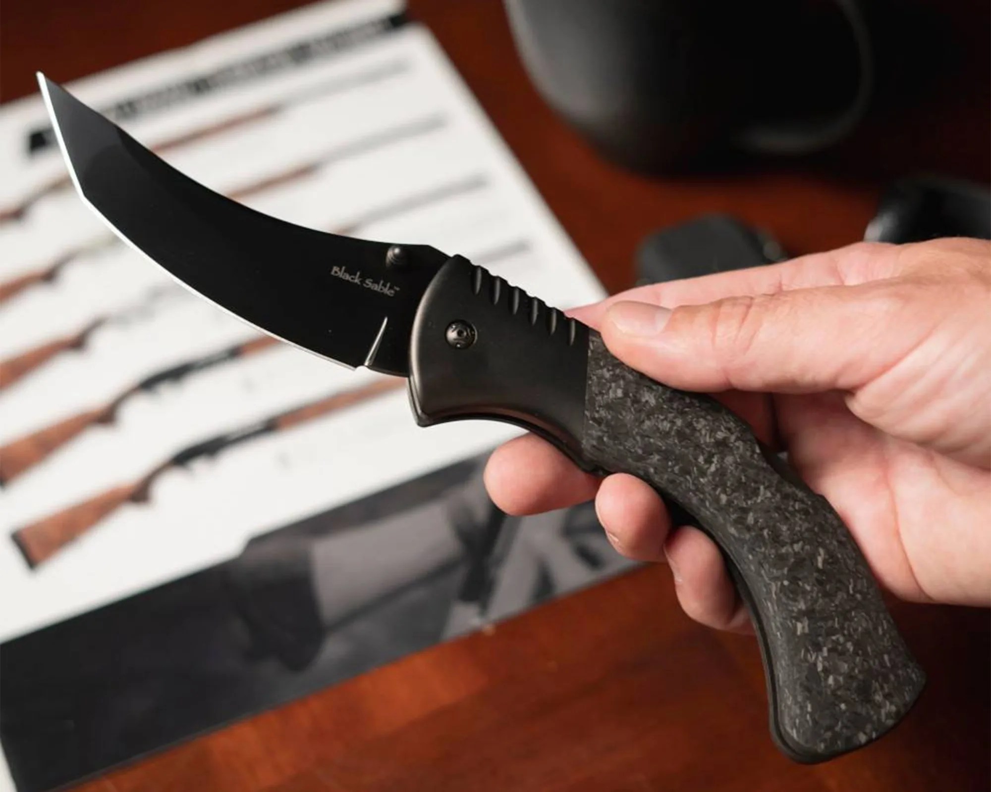 Cold Steel Black Sable EDC Knife in hand above coffee mug on wood desk with watch, glasses, pen, etc.