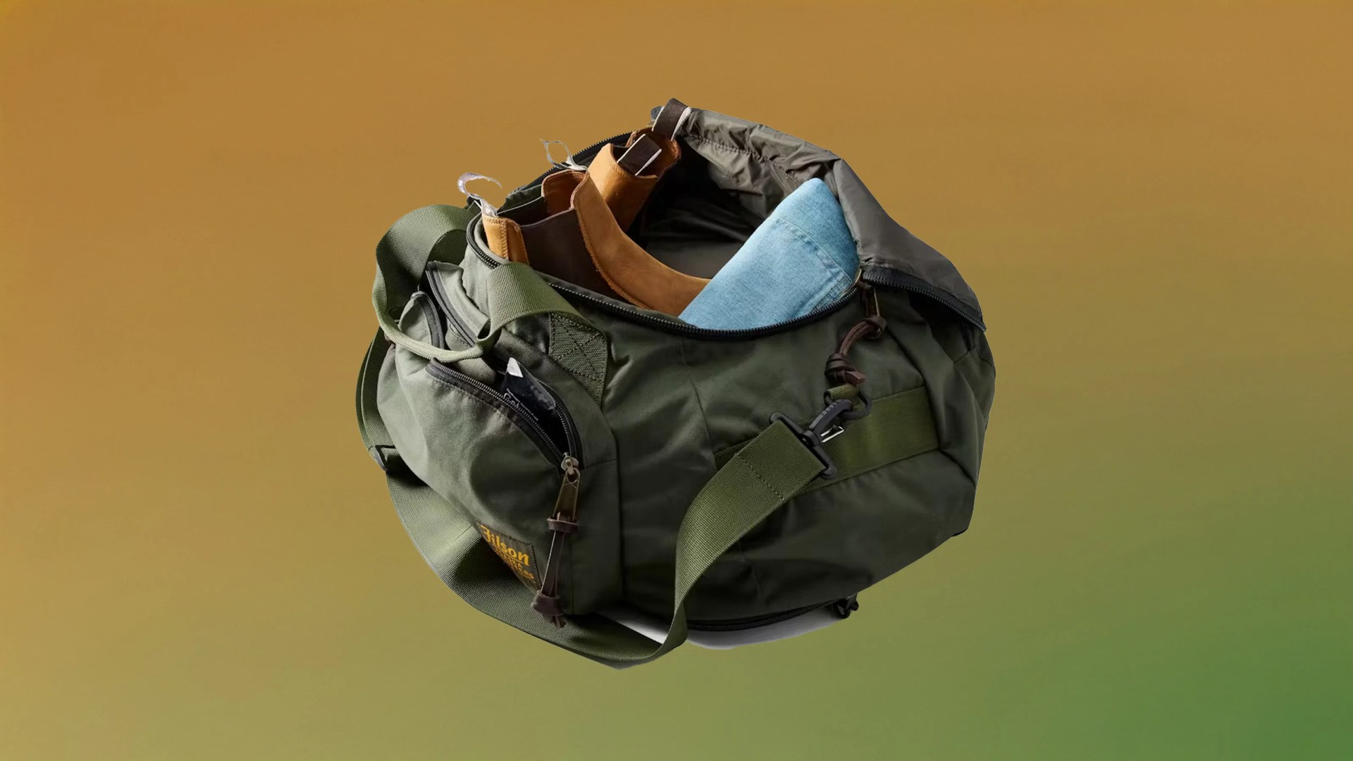 The Filson Travel Duffle shown open with jeans and boots packed inside against a gradient green and brown background