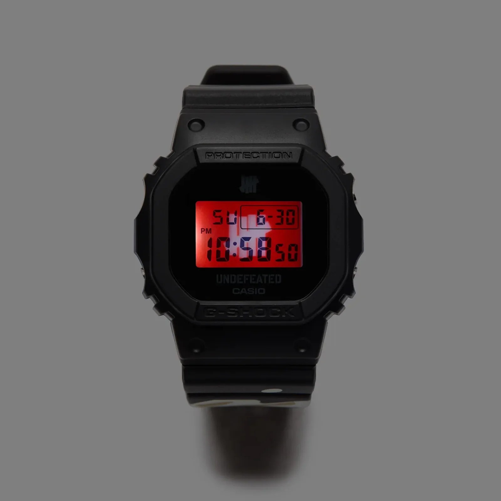 The G-Shock and 'Undefeated' DWE5600UD with its red light illuminated against a dark grey background