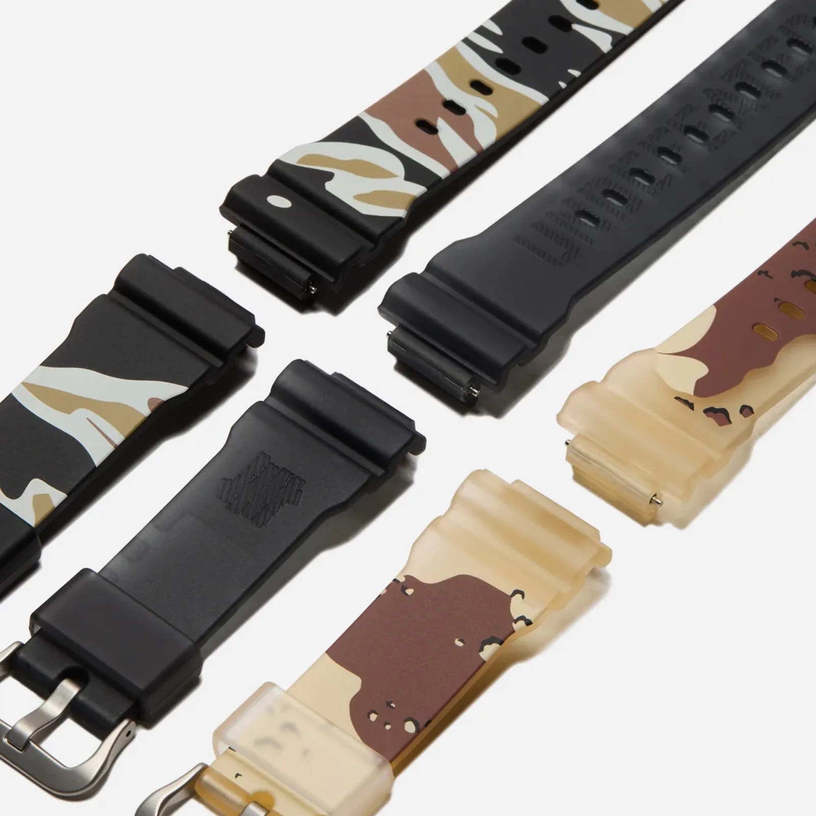 The three straps included with the he G-Shock and 'Undefeated' DWE5600UD collab watch shown lying flat on at an angle lined up in a row. One strap is black in the middle, and the other two are camo patterns flanking the black strap on either side. 