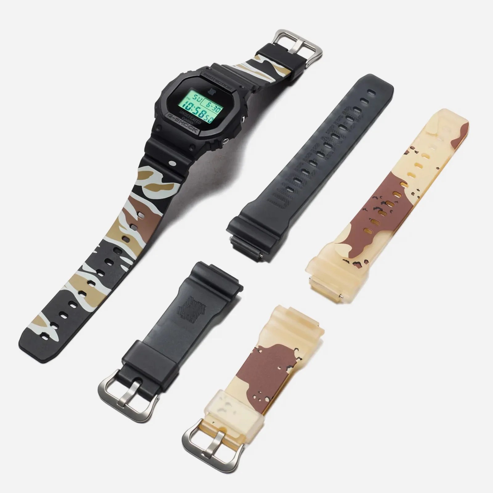 The Undefeated x G-Shock DWE5600 shown on a camo strap in a line next to the two other straps included with the watch.