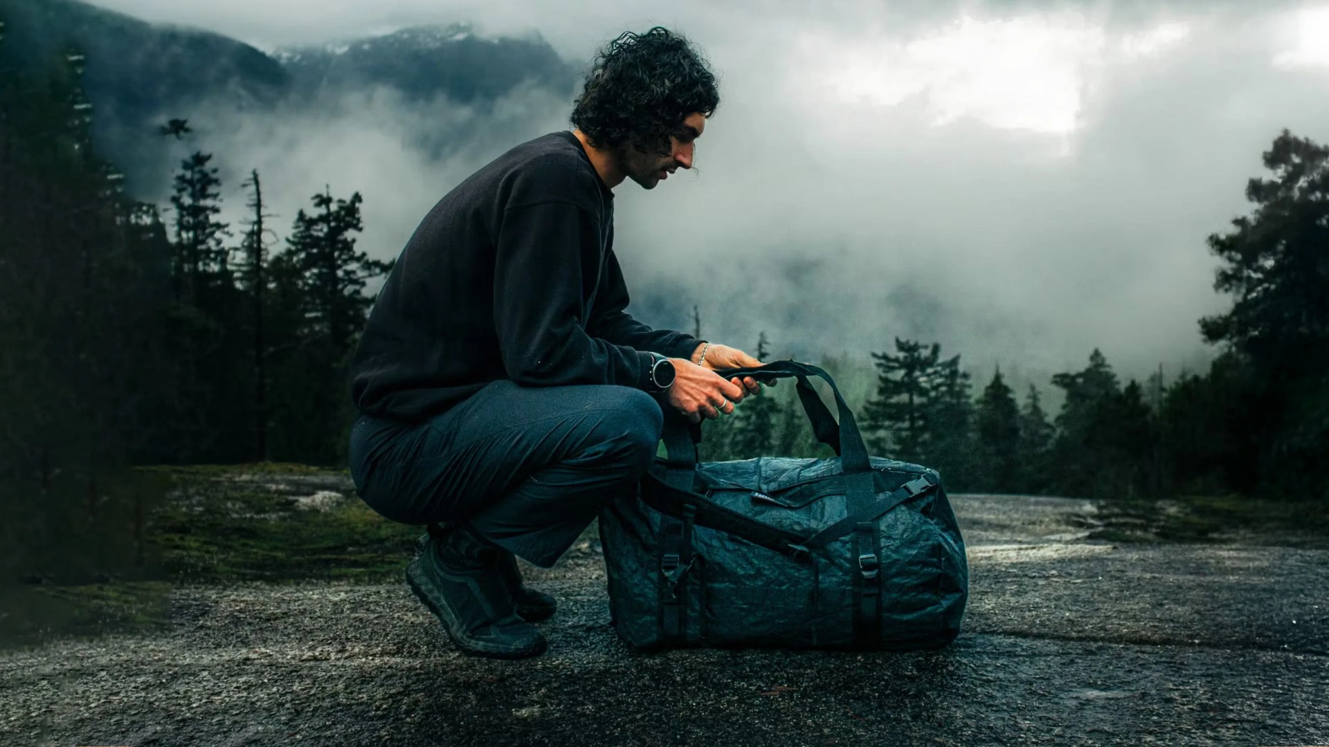 A man squatting over a Norda's Toolbox Dyneema Duffel Bag in black sitting on a asphalt surface with pine greens and clouds in the background