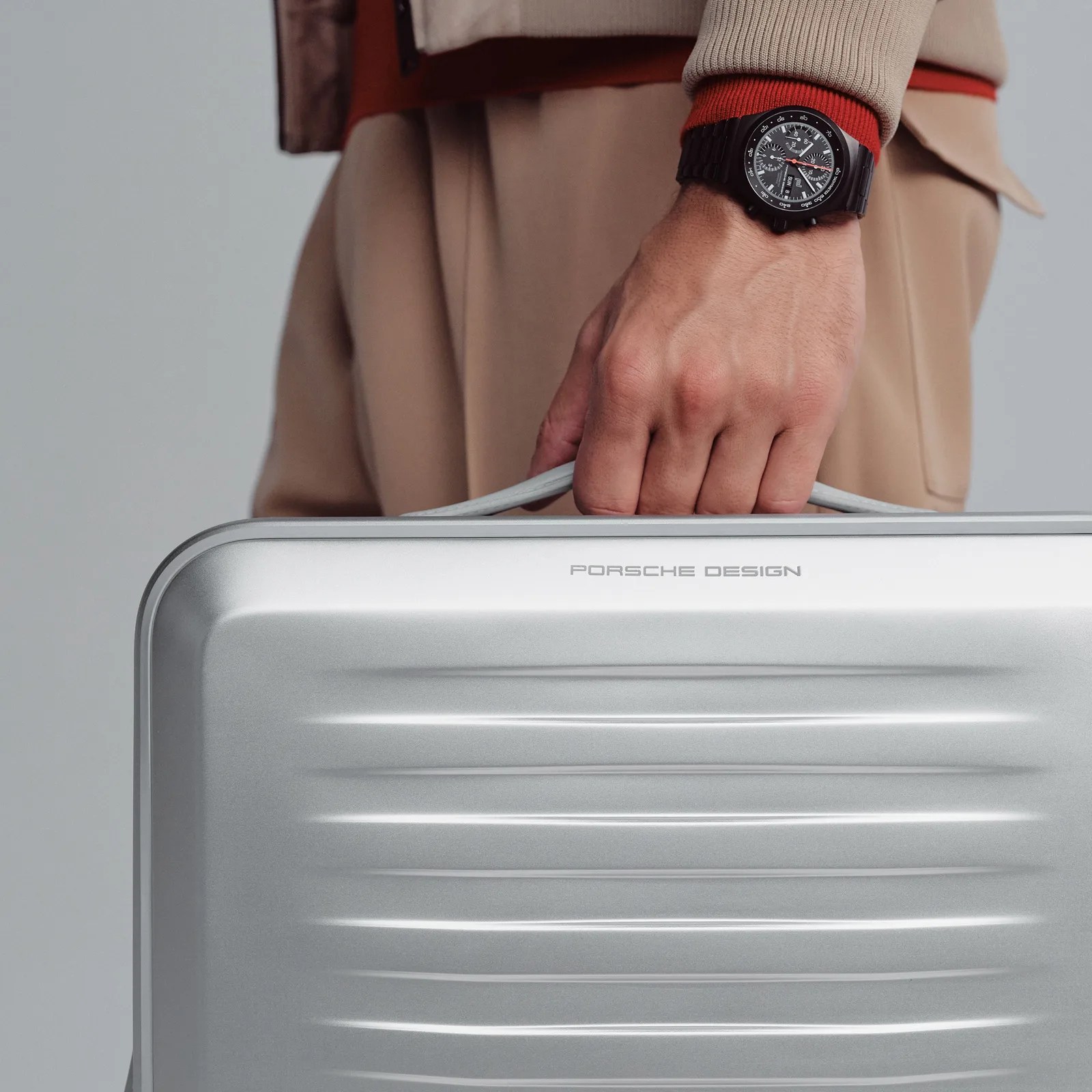 A man's hand is shown holding the top handle of the Porsche Design Roadster aluminum suitcase.