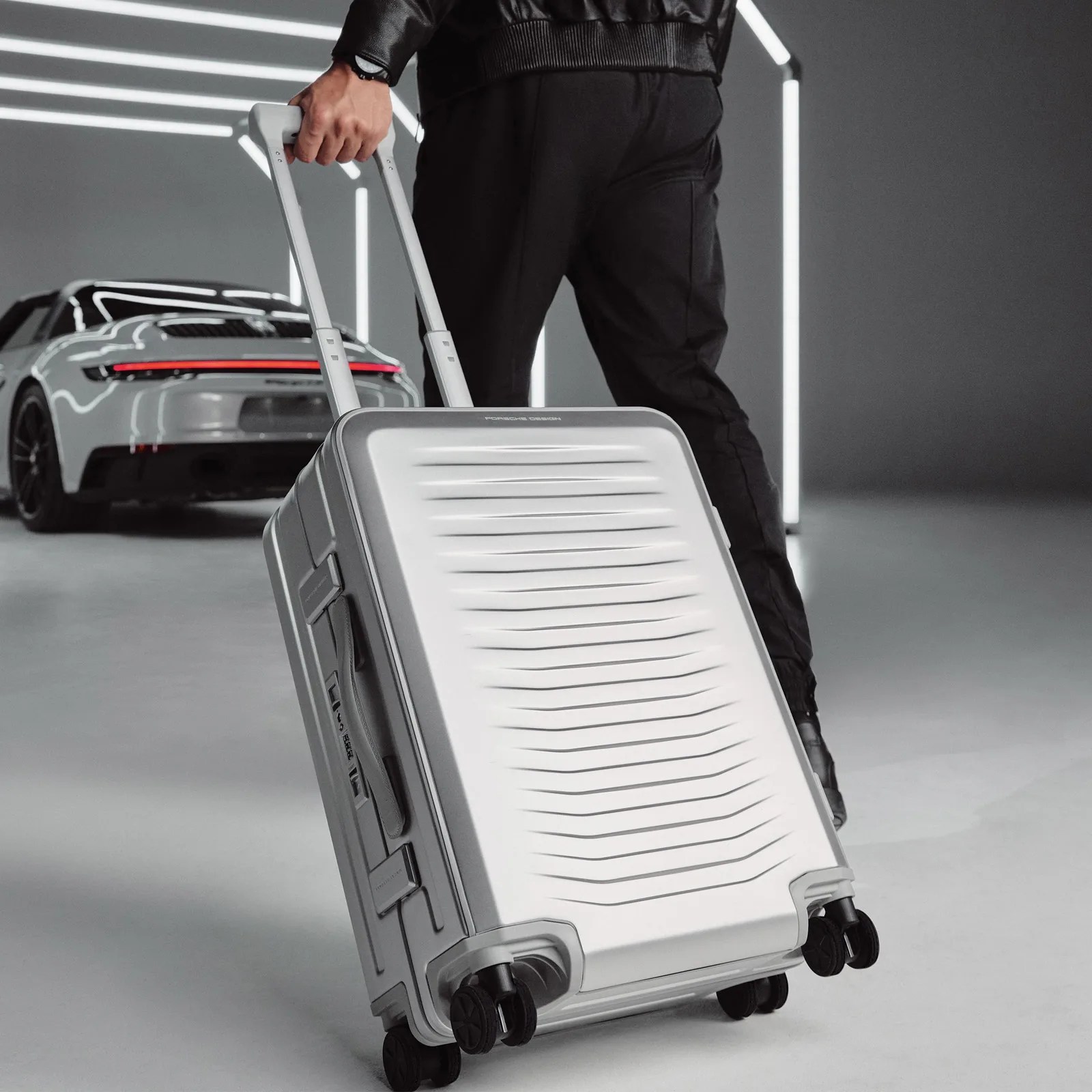 A man's hand is shown rolling the Porsche Design Roadster aluminum suitcase with a Porsche car in the background. 