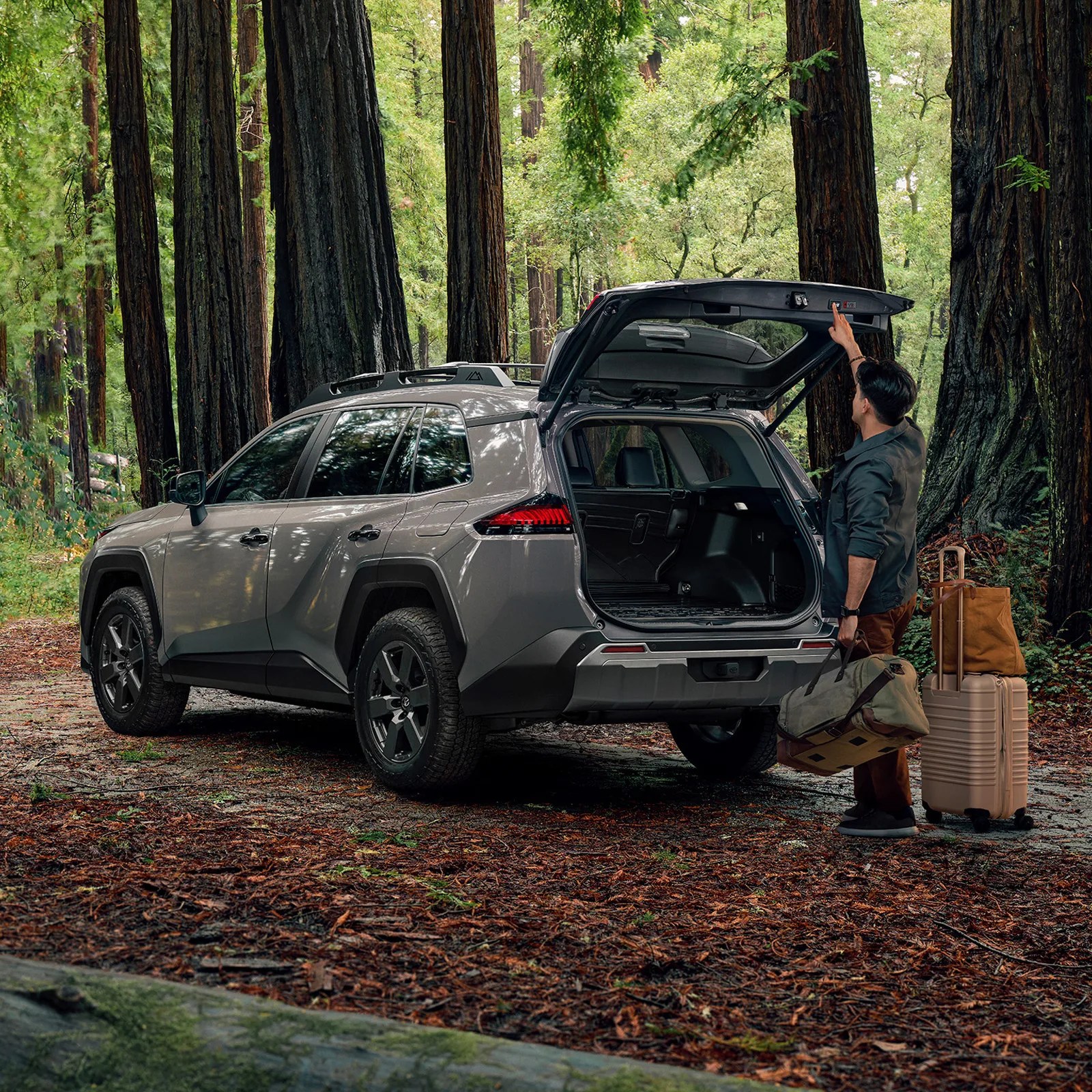 A man shown closing the rear hatch of the 2026 Toyota RAV4 Woodland Edition holding a duffle bag and standing next to a roller suitcase. The car is parked on a leaf-covered ground surrounded by trees. 