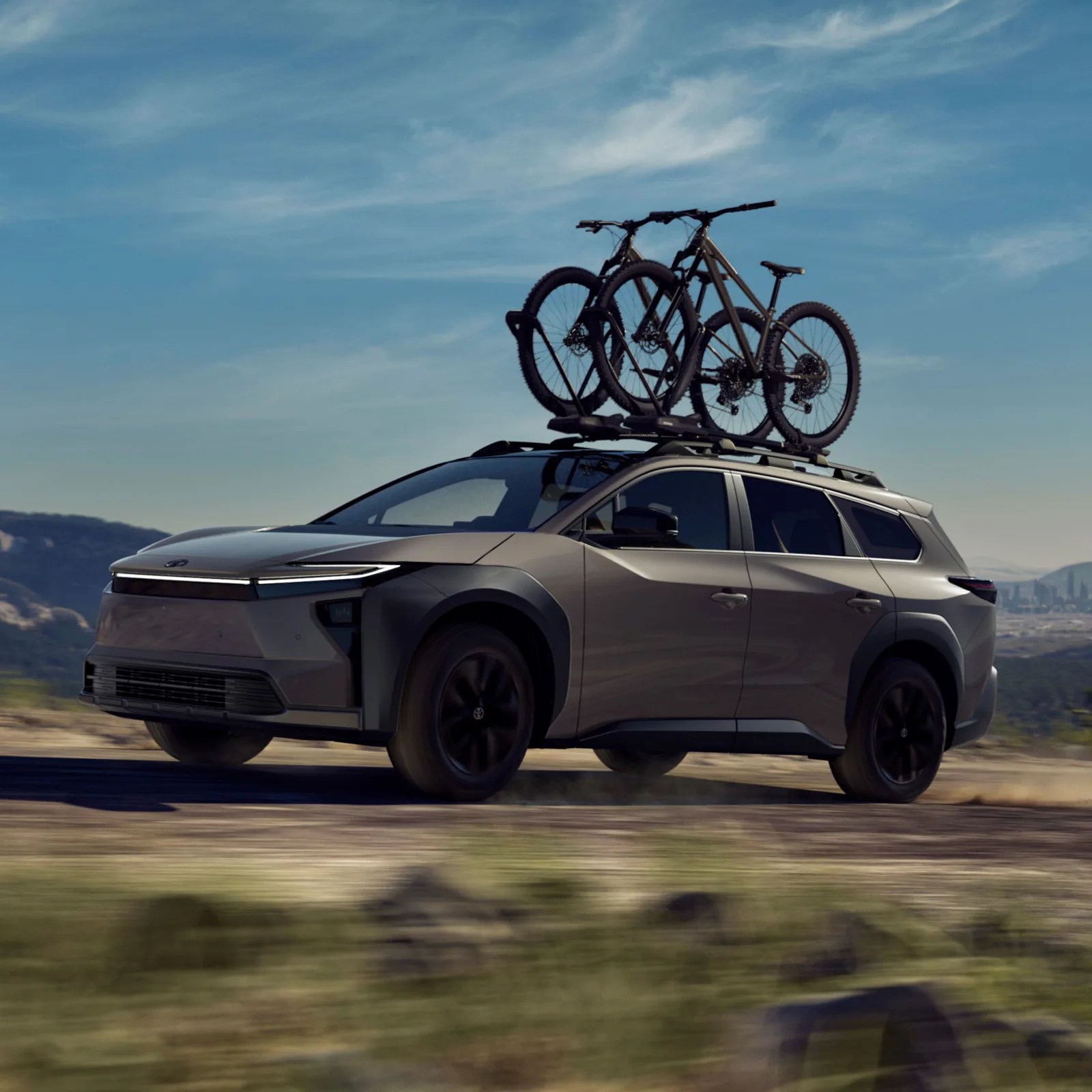The Toyota bZ Woodland EV in a light brown color shown driving fast down a dirt road at an angle with a roof rack carrying bikes.