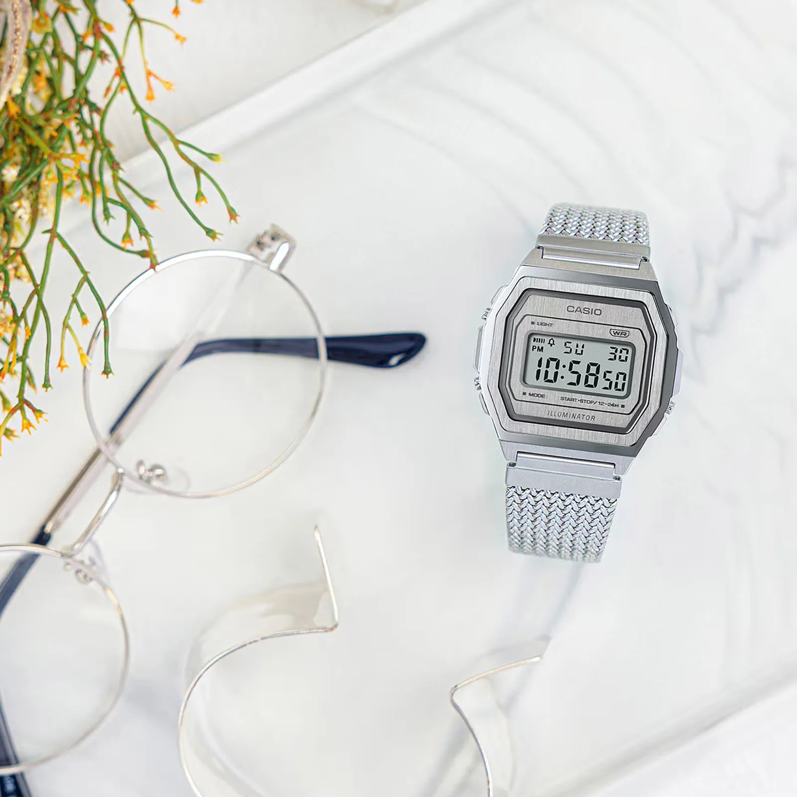 A Casio A1000MA-7VT shown sitting on a marbled white table next to a pair of eyeglasses with circular frames and a plant