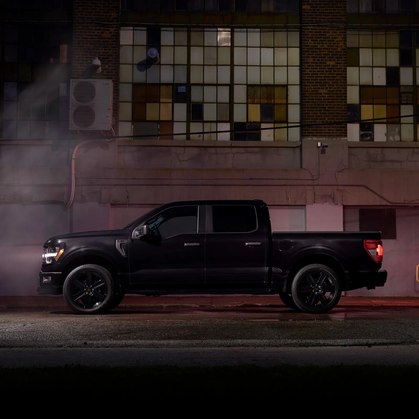A profile view of the 2025 Ford F-150 Lobo in black shown with its headlights on against a grey warehouse wall