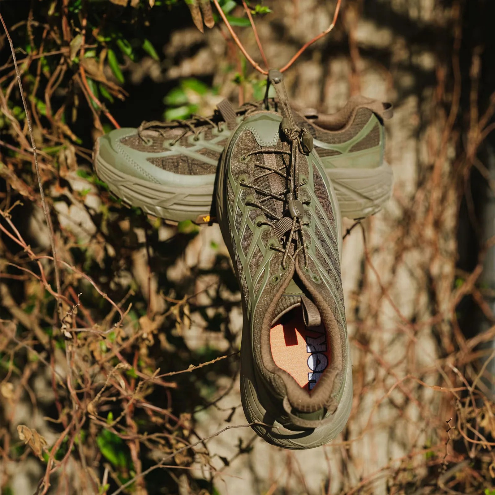A pair of Hoka Mafate Three2 Grid JP in "antique olive and eucalyptus" hanging from a branch in a wooden background. 