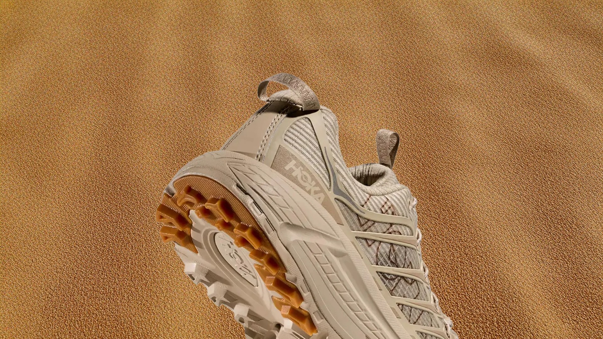 The heel of a Hoka Mafate Three2 Grid JP Sneaker in Sand / Linen against a sandy textured background