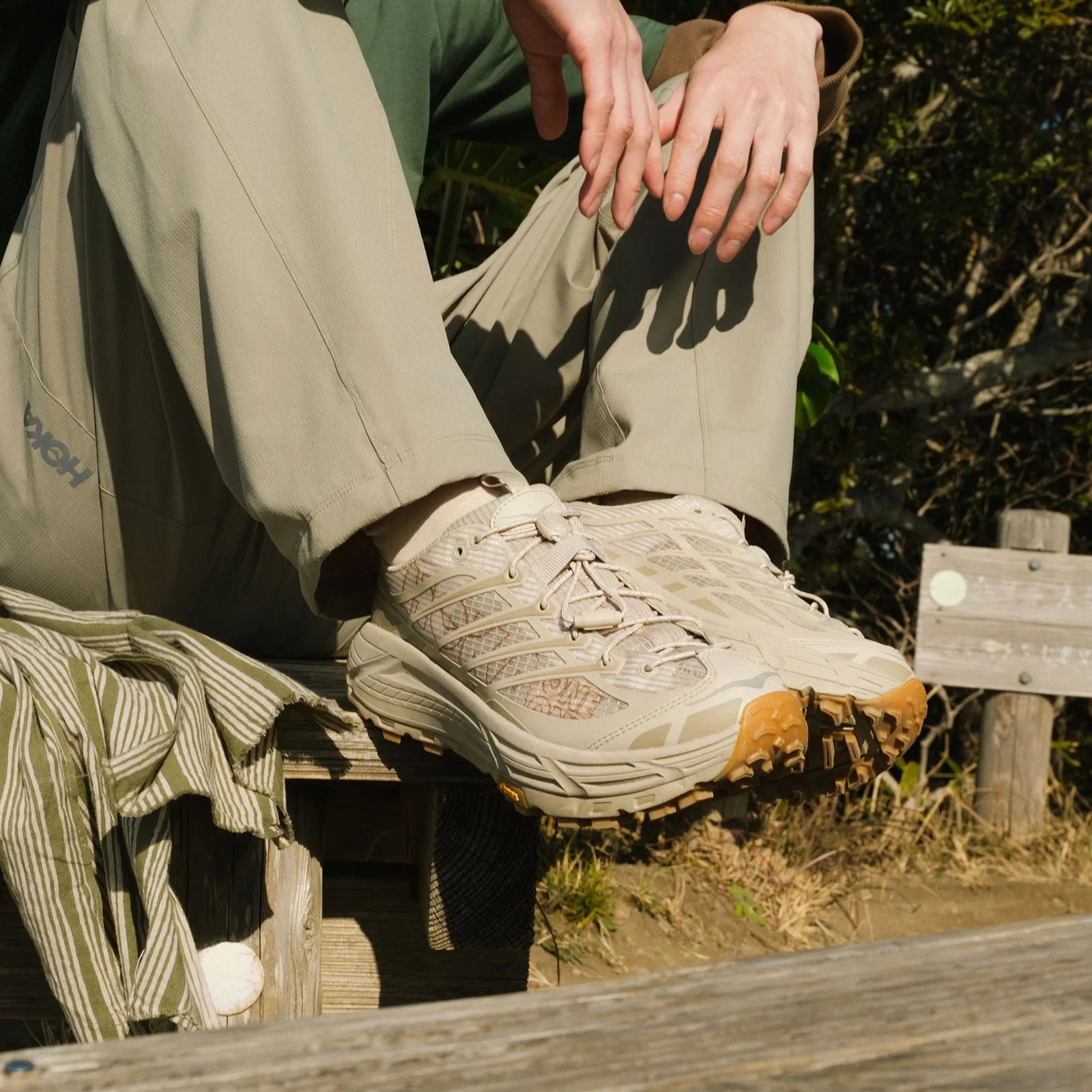 A man wearing a pair of Hoka Mafate Three2 Grid JP in "Raw Linen / Sand" sitting down on a wooden bench. 