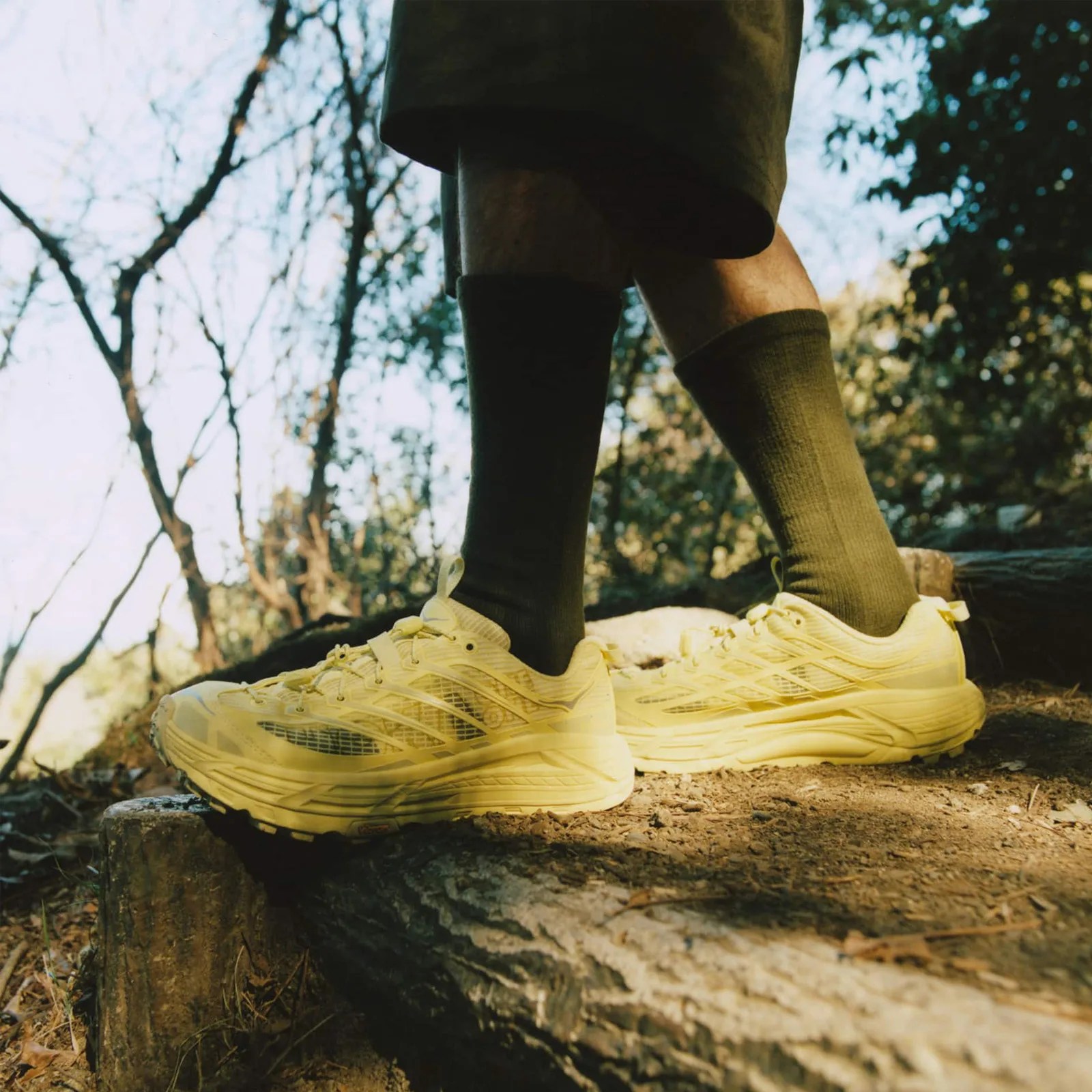 A person wearing high olive green ankle socks wearing a pair of Hoka Mafate Three2 Grid JP in "Yuzu / Cardamom" walking on trail shown from the knees down. 