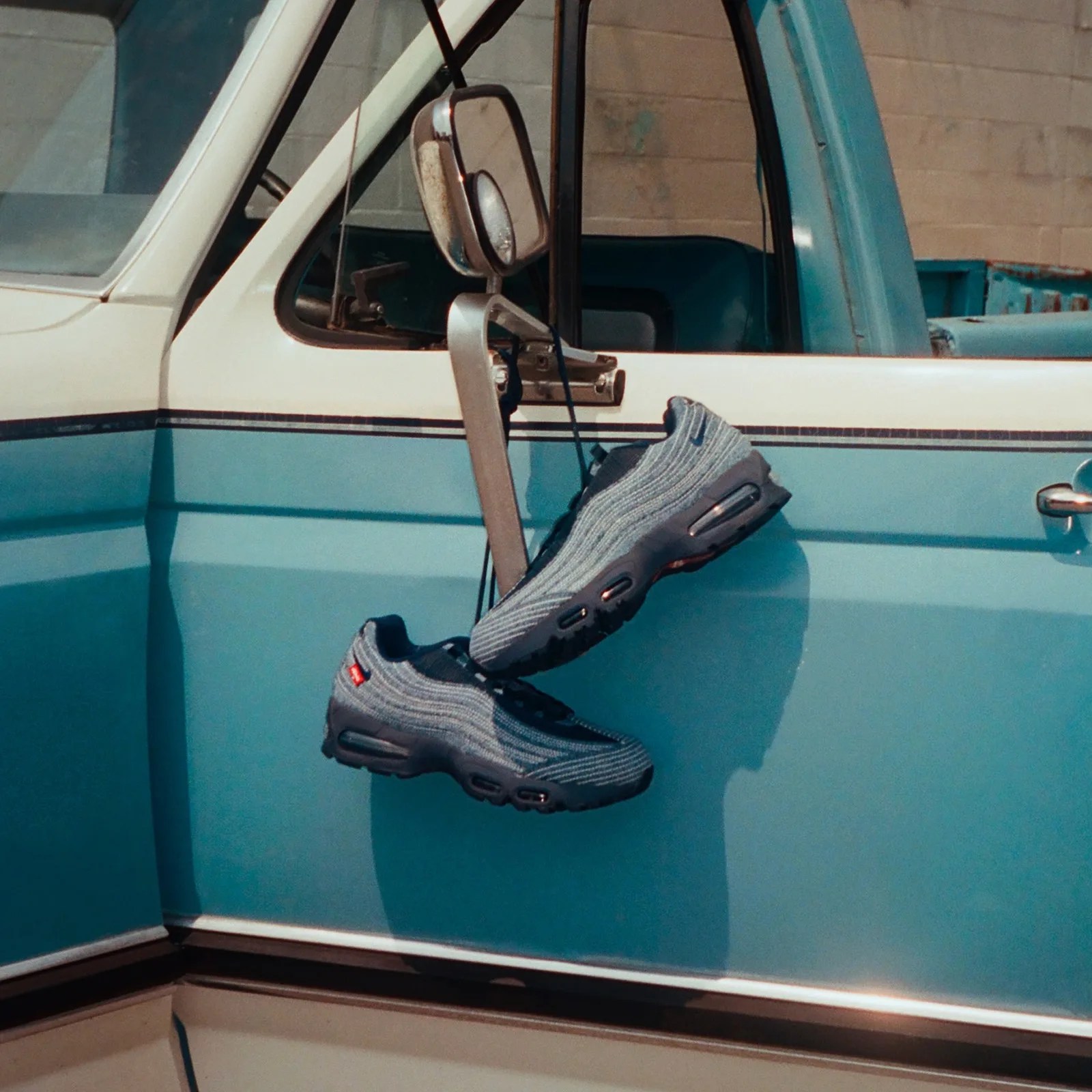 A pair of blue Nike x Levi's Air Max 95's hanging over a side view mirror of a vintage truck colored in white or teal
