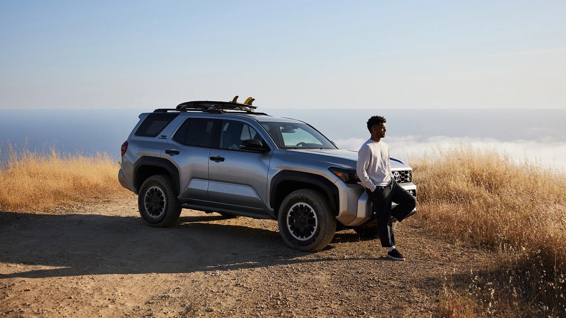 Surfer hunter jones leans against a silver 2025 Toyota 4Runner TRD Off-road with a surfboard strapped to the roof on a backdrop of the pacific ocean