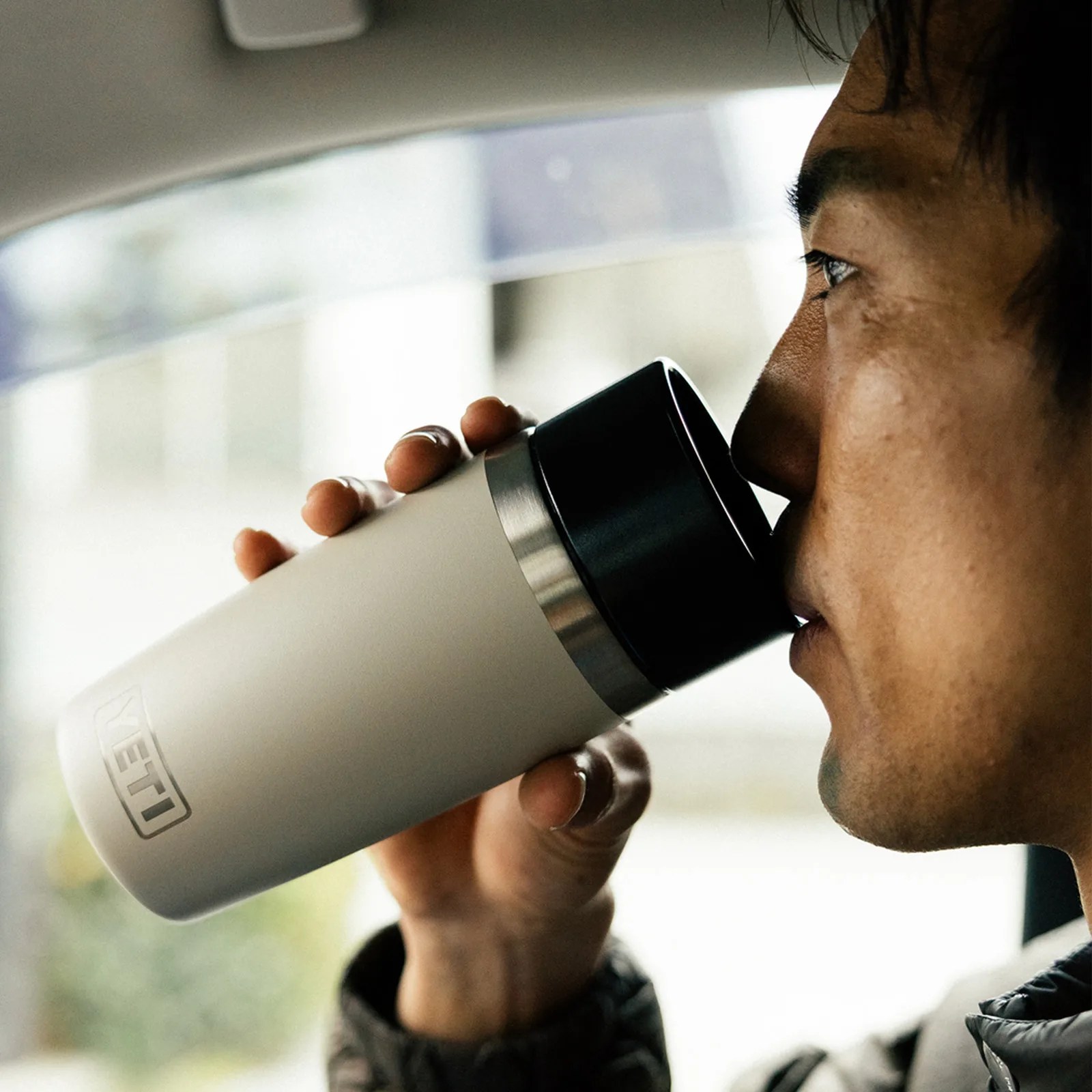 A close-up of a man drinking out of a Yeti Travel bottle in white while sitting in a car