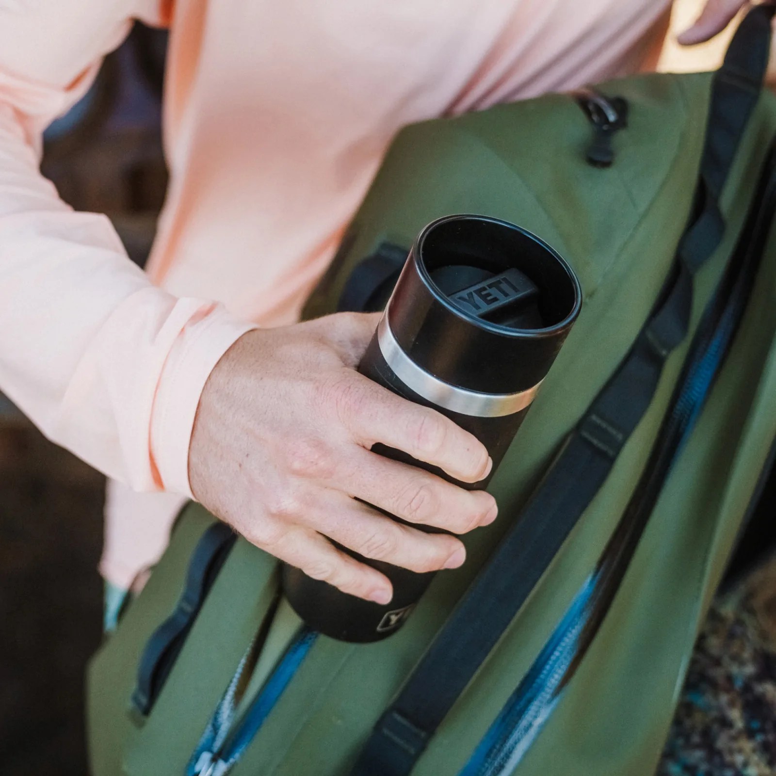A man wearing a pink long sleeve shirt shown sliding a black Yeti Travel bottle into a green bag