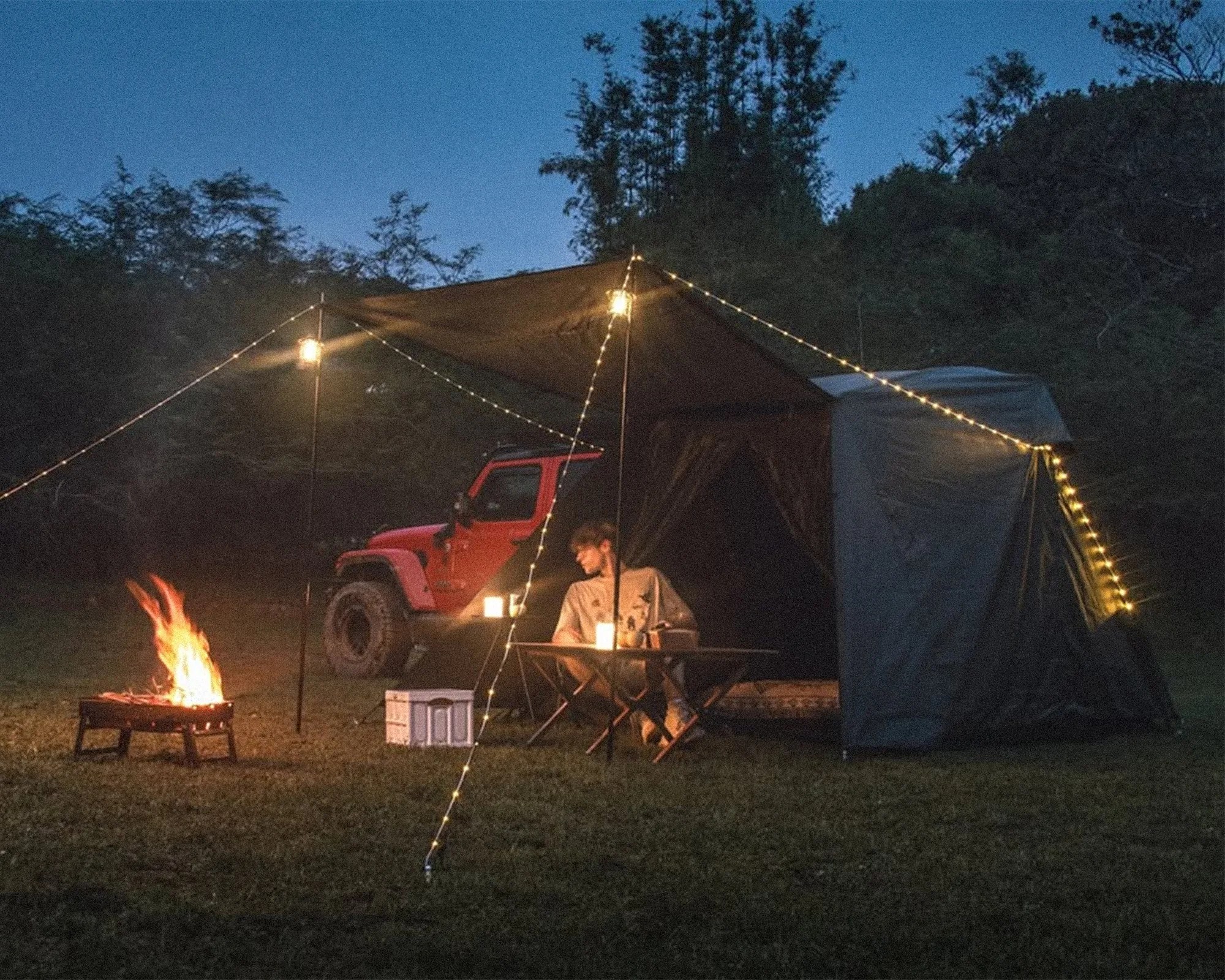 Altara Dual Awning Modular Camping Tent on evening with Jeep, camper, camp table, cooler, fire pit, lights in forest clearing
