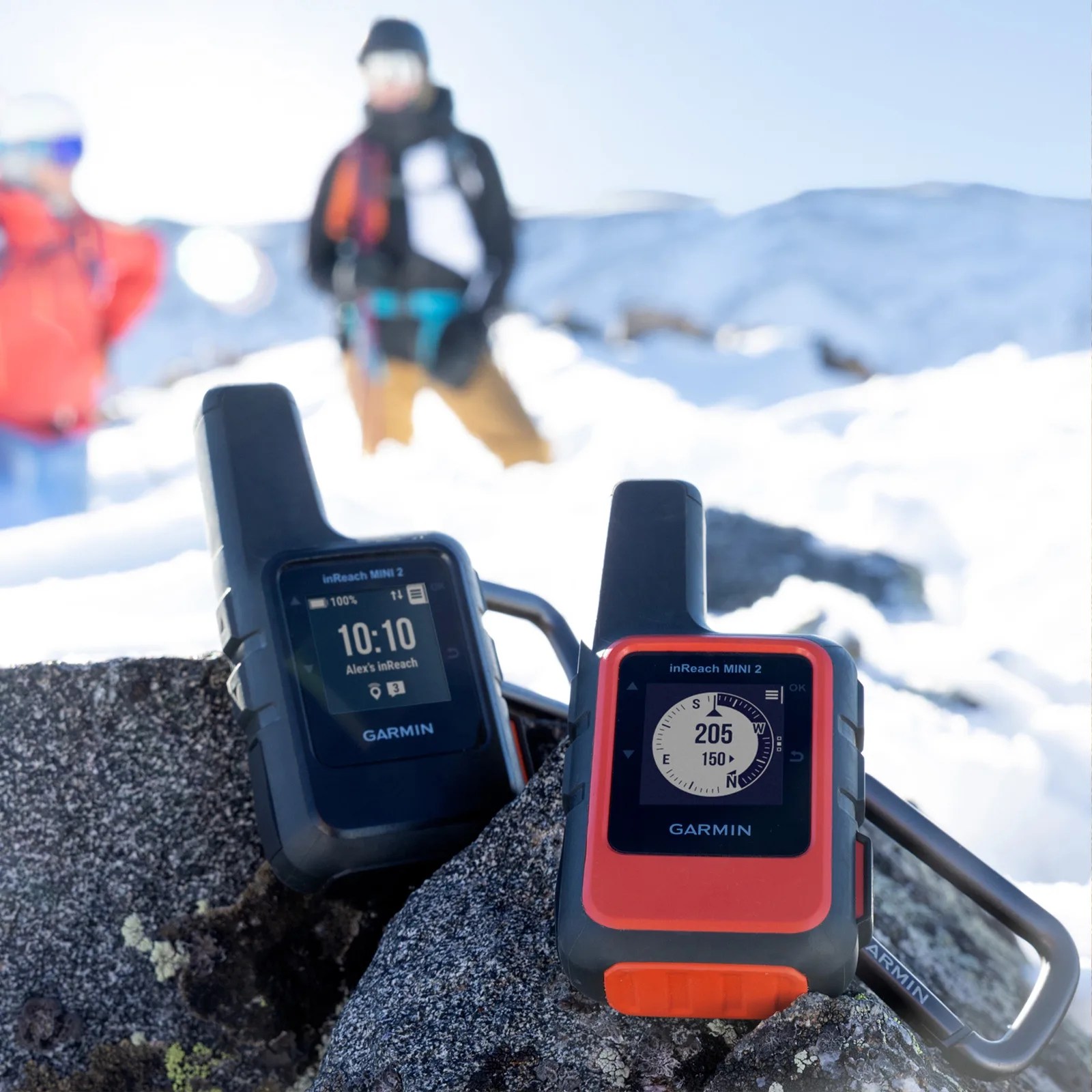 A pair of Garmin inReach satellite messenger devices sitting on a rock with two skiers on a snowy mountain in the background