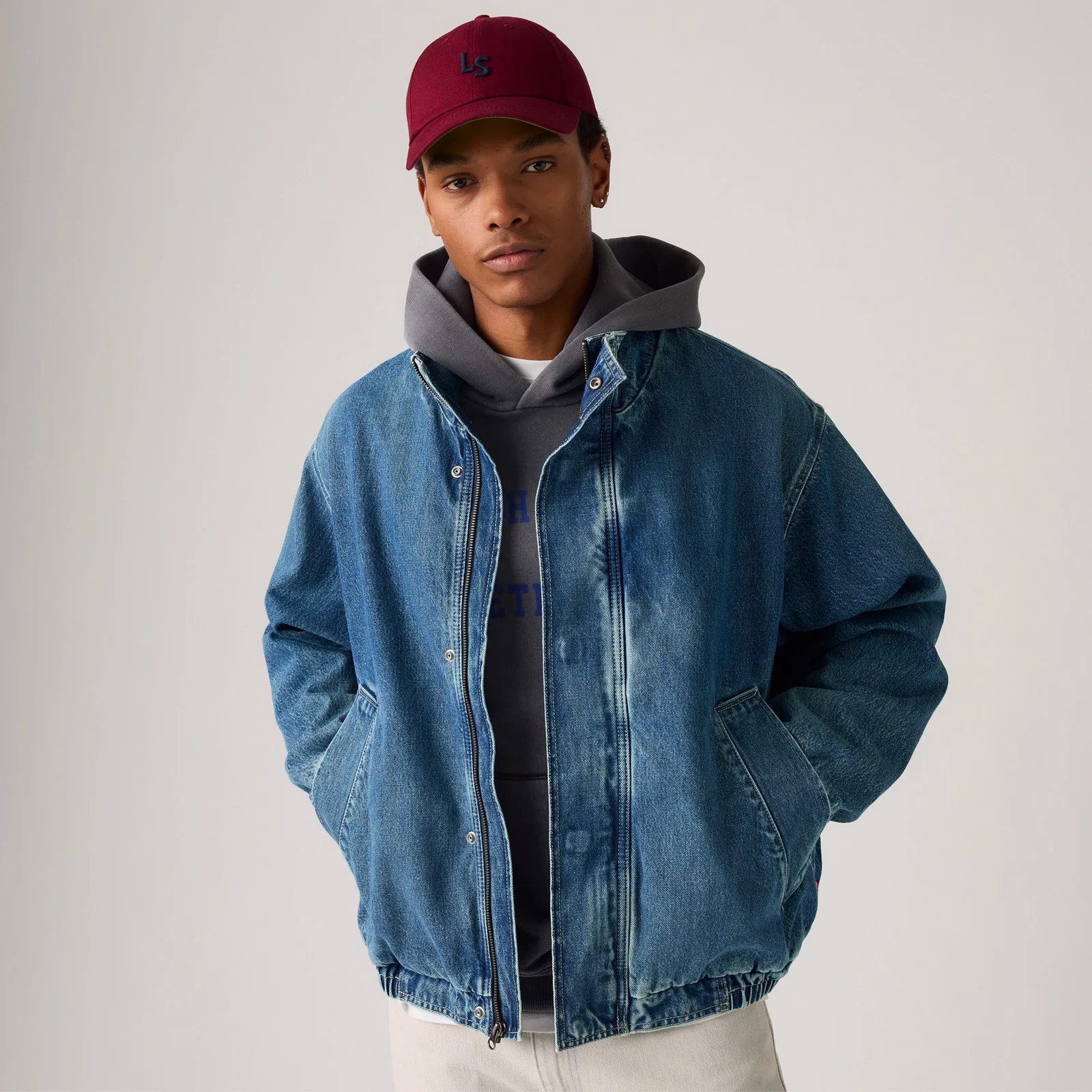 A male model is shown wearing the Levi's Highland Park Denim Jacket over a hooded sweatshirt, looking straight ahead with his hands in his pocket