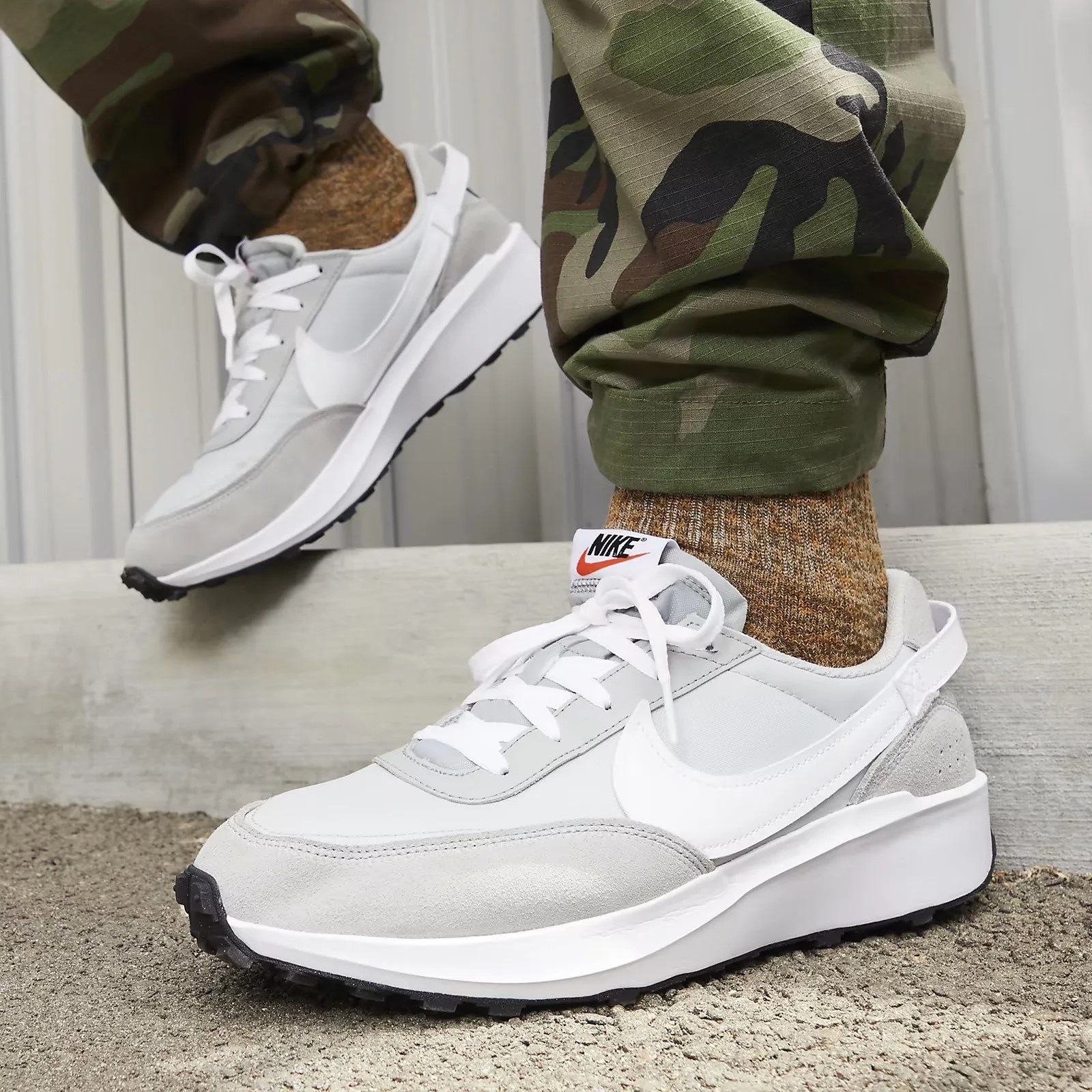 A man wearing a pair of grey and white Nike Waffle Debut sneakers is shown from the ankles down. The man is also wearing camouflage pants