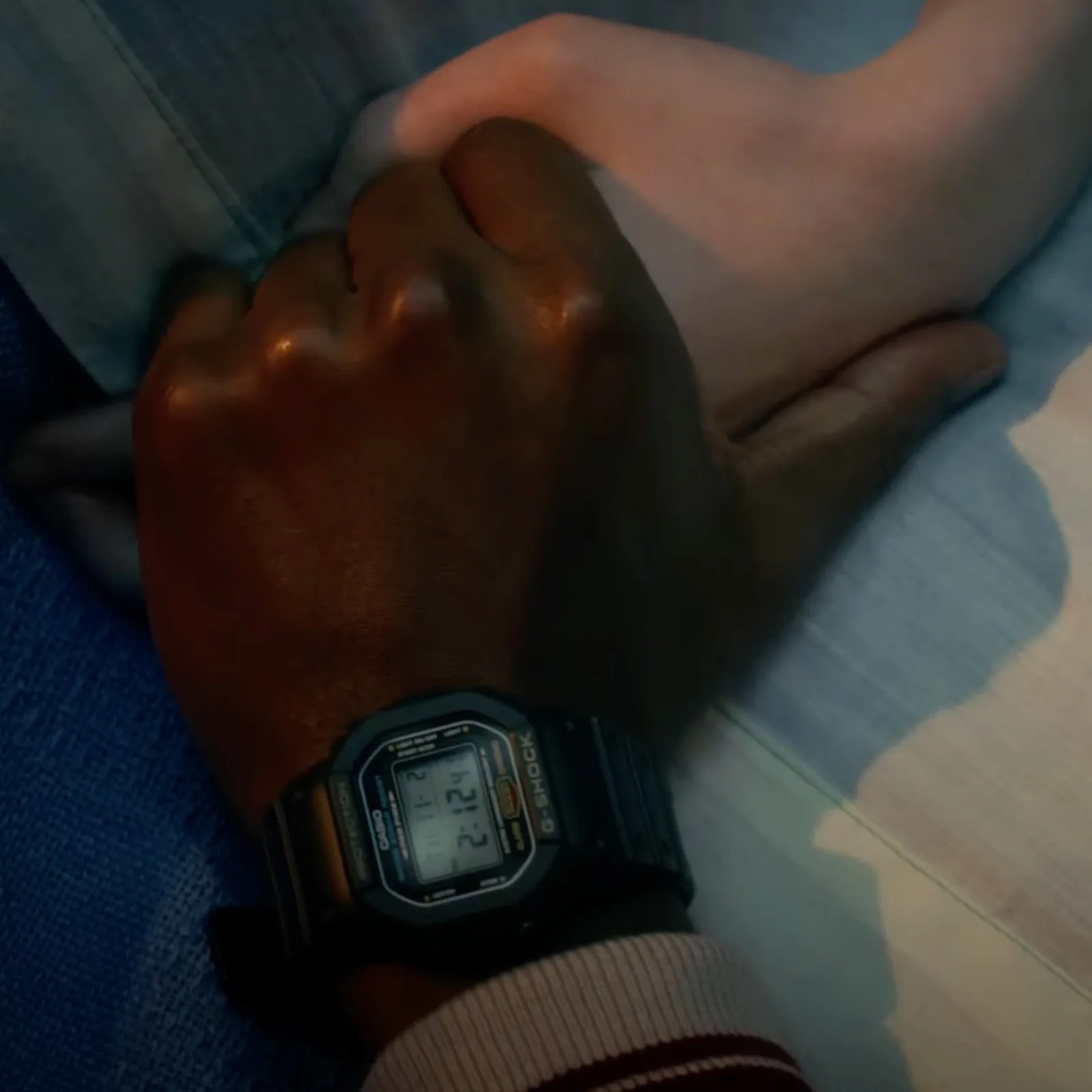 A screenshot from the Netflix TV show Stranger Things showing the wrist of Lucas Sinclair wearing a Casio G-Shock DW5600E while holding the hand of another character lying in a hospital bed