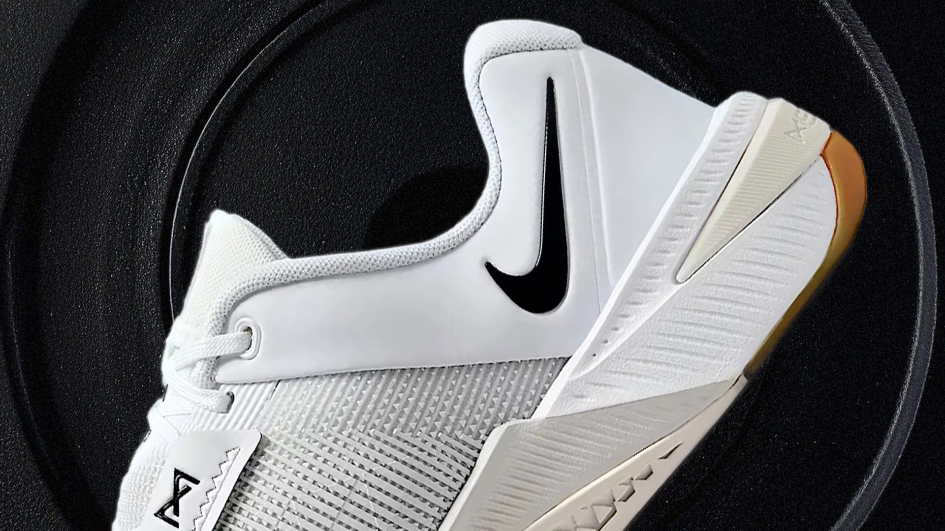 A close up of the side of the Nike Metcon 10 gym shoe in white against what appears to be a metal weight lifting plate